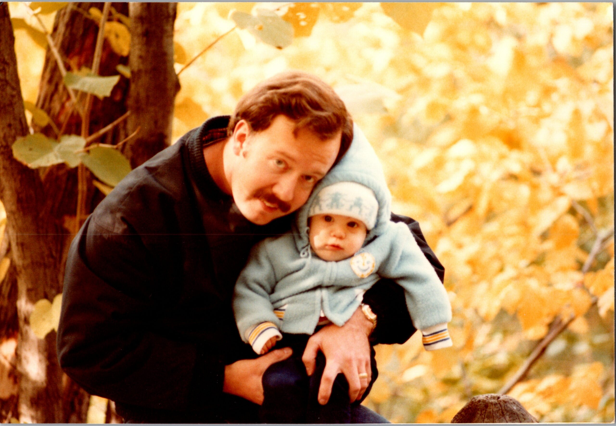 Man with a mustache holding a baby in a blue outfit, outdoors among autumn leaves in a park.