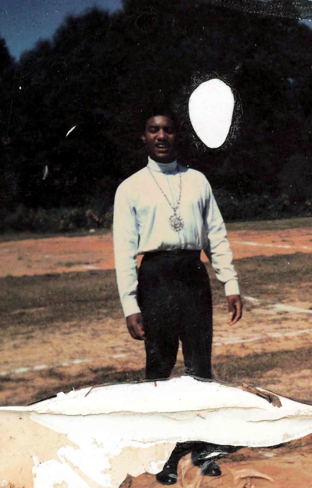 Person standing outdoors in a light shirt and dark pants, wearing a long necklace with a large pendant, on a grassy field with trees behind.