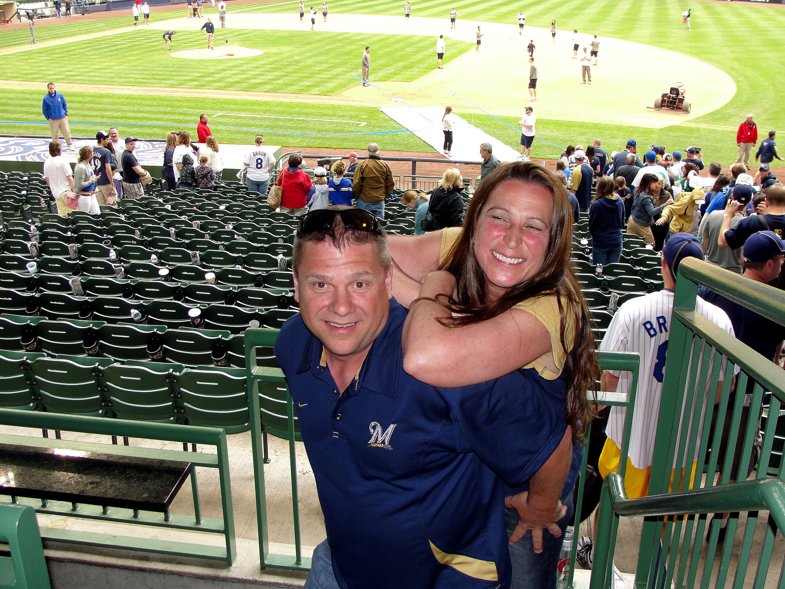 Smiling man and woman hug at the railing in a crowded baseball stadium, with the field and players visible in the background.