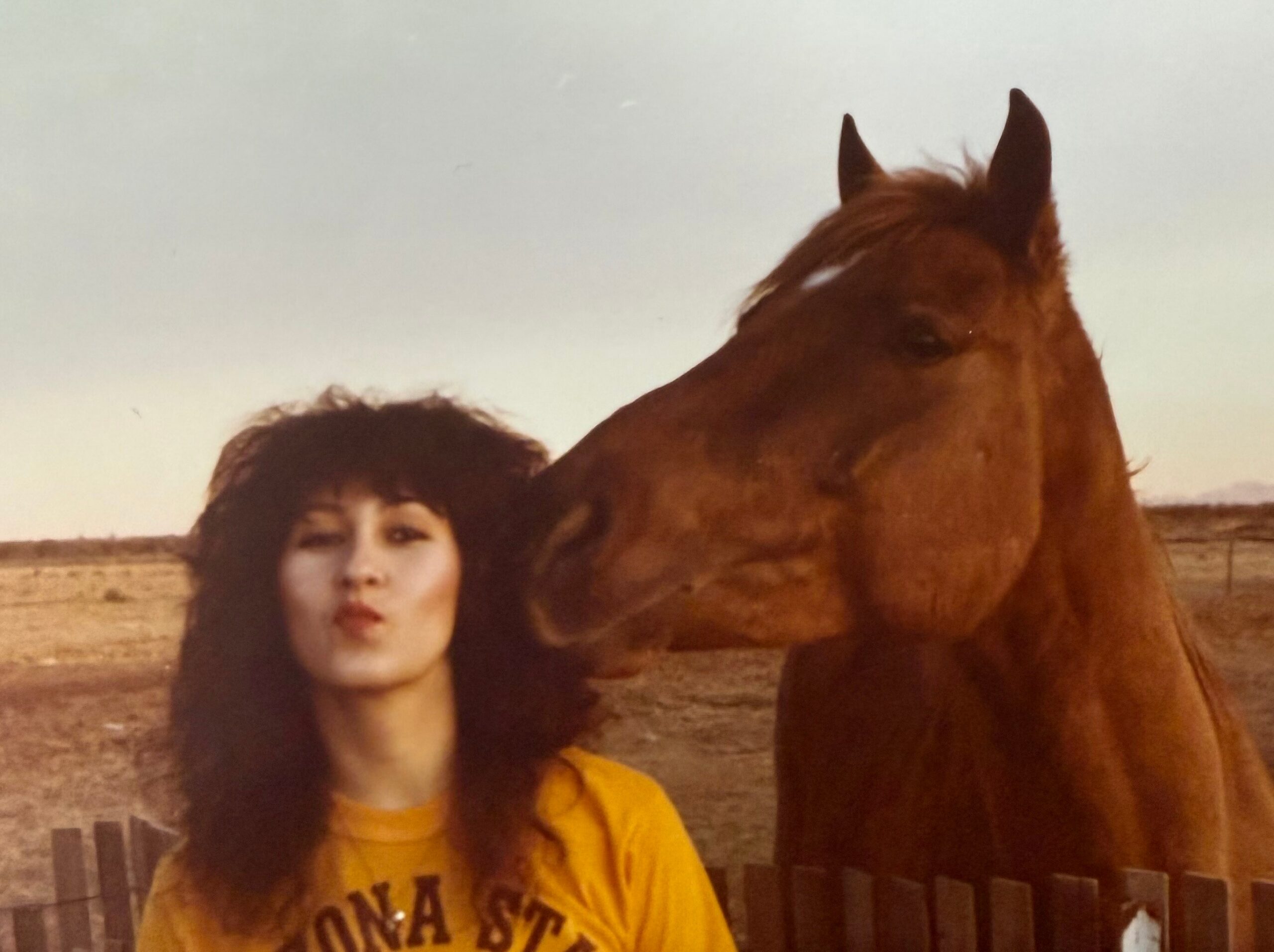 A woman with long dark hair wearing a yellow shirt stands beside a large brown horse outdoors near a wooden fence.