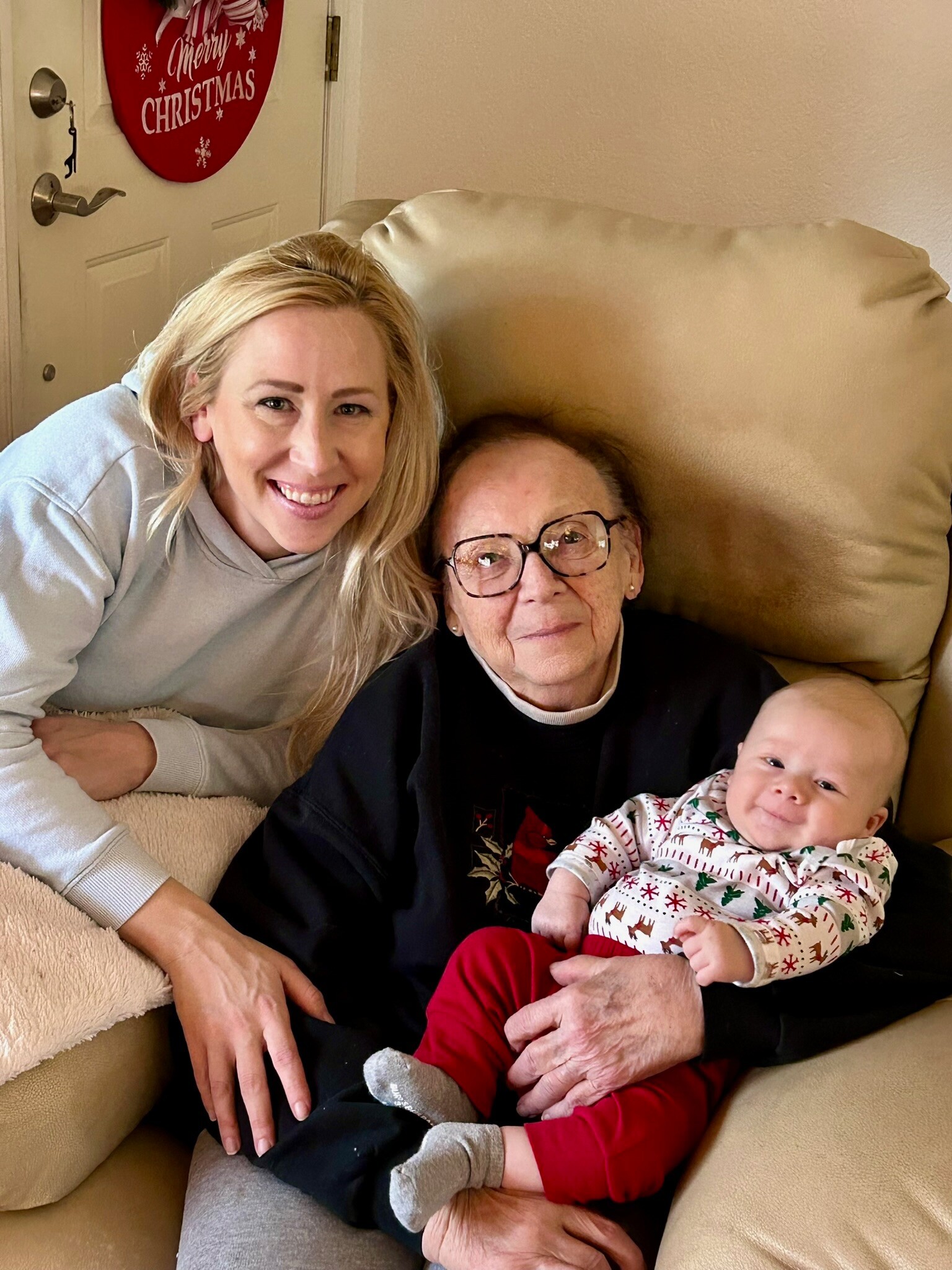 A smiling woman and an elderly woman holding a baby on a beige couch near a Christmas door banner.