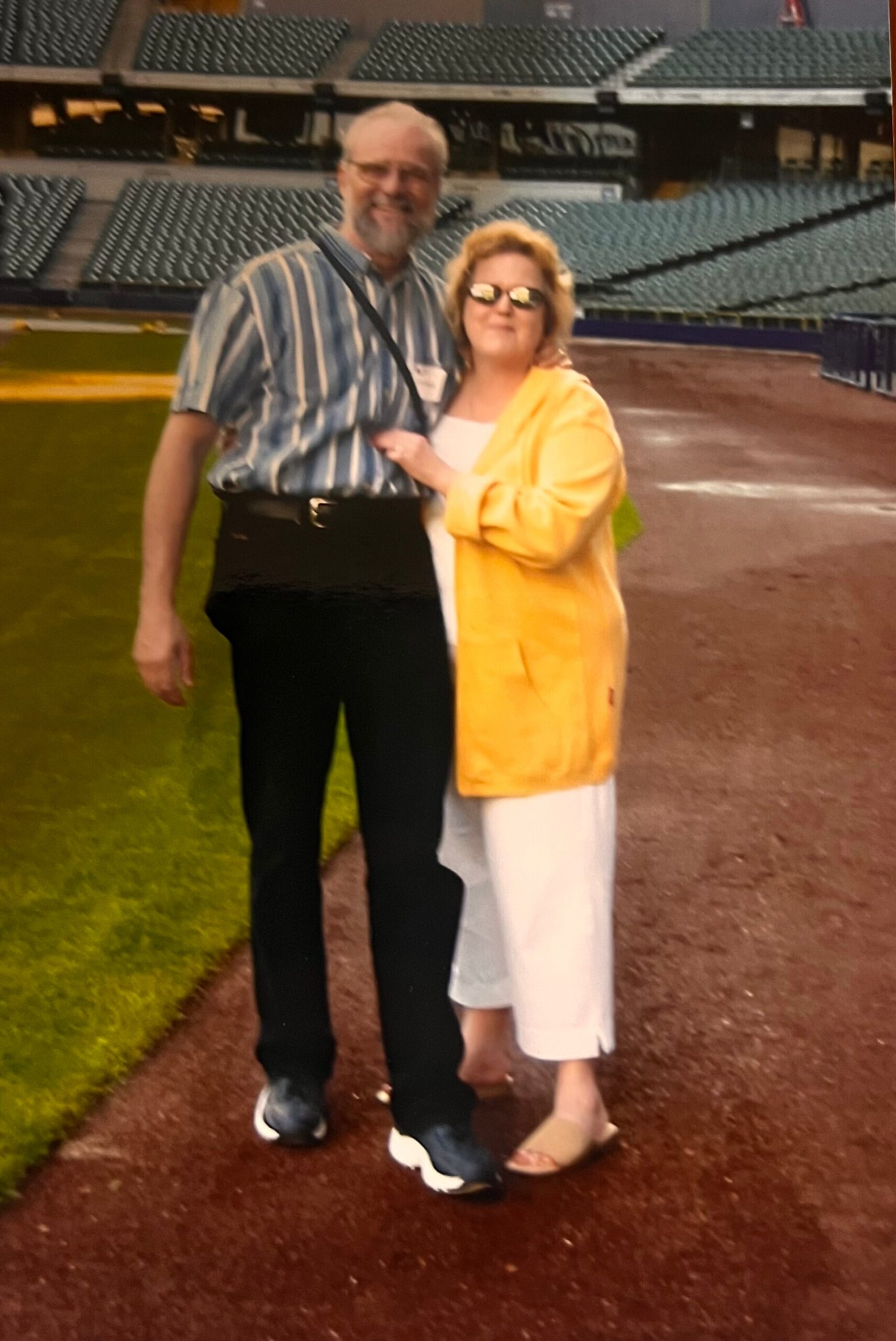 Smiling couple standing arm-in-arm on a baseball field with empty stadium seats in the background.