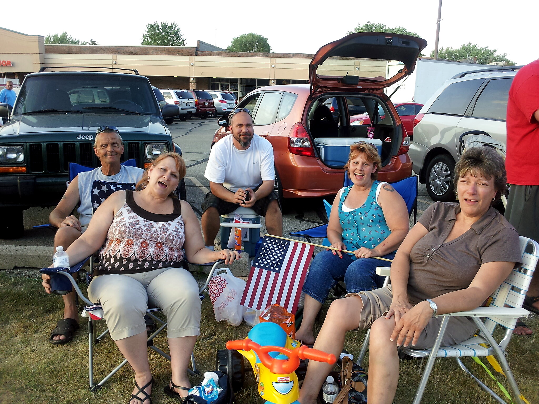 Group of adults sitting in folding chairs in a parking lot, with an open car trunk behind them and an American flag nearby.