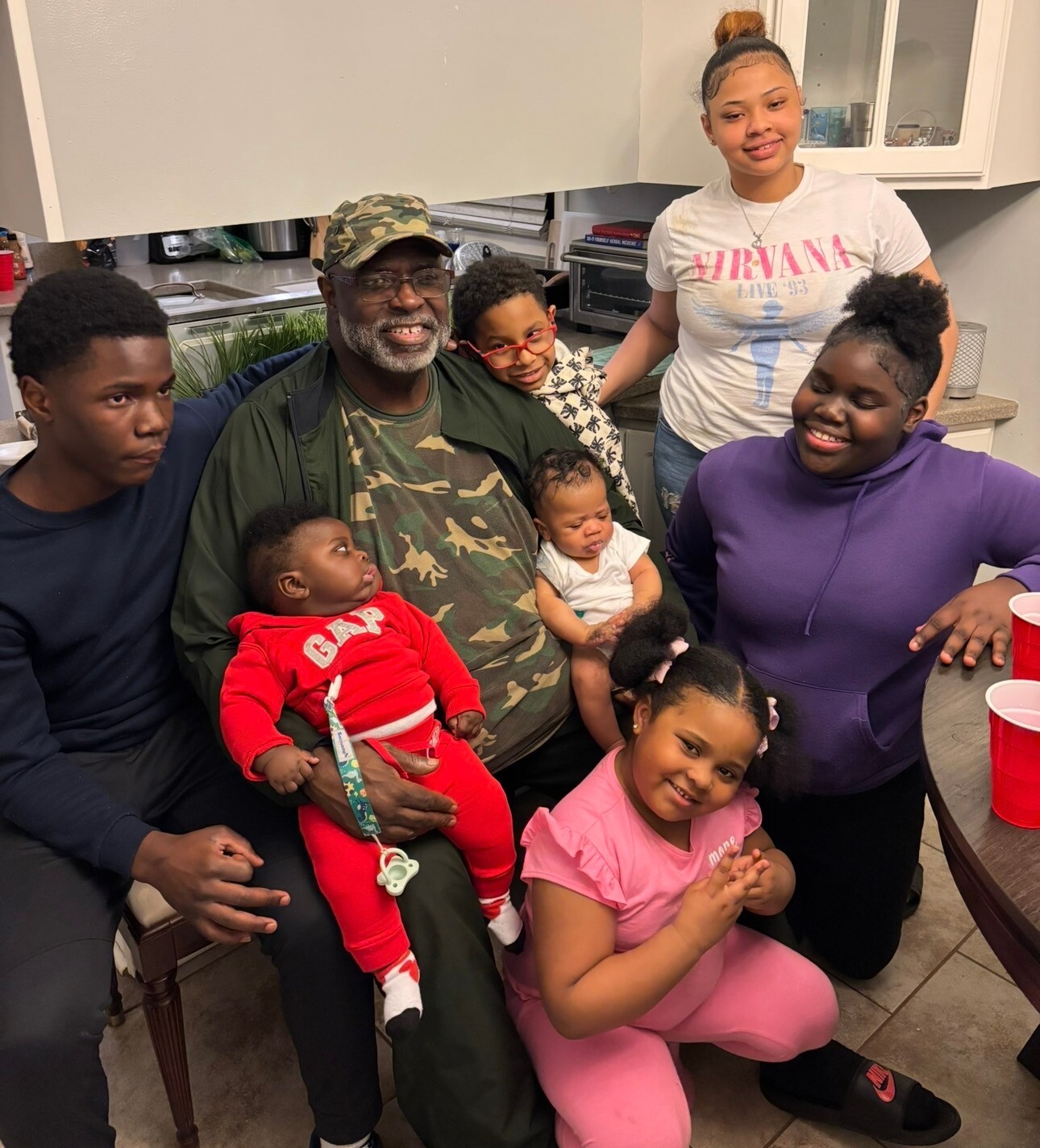 Family portrait in a kitchen: man in camo holds a baby while several children and adults smile around him.