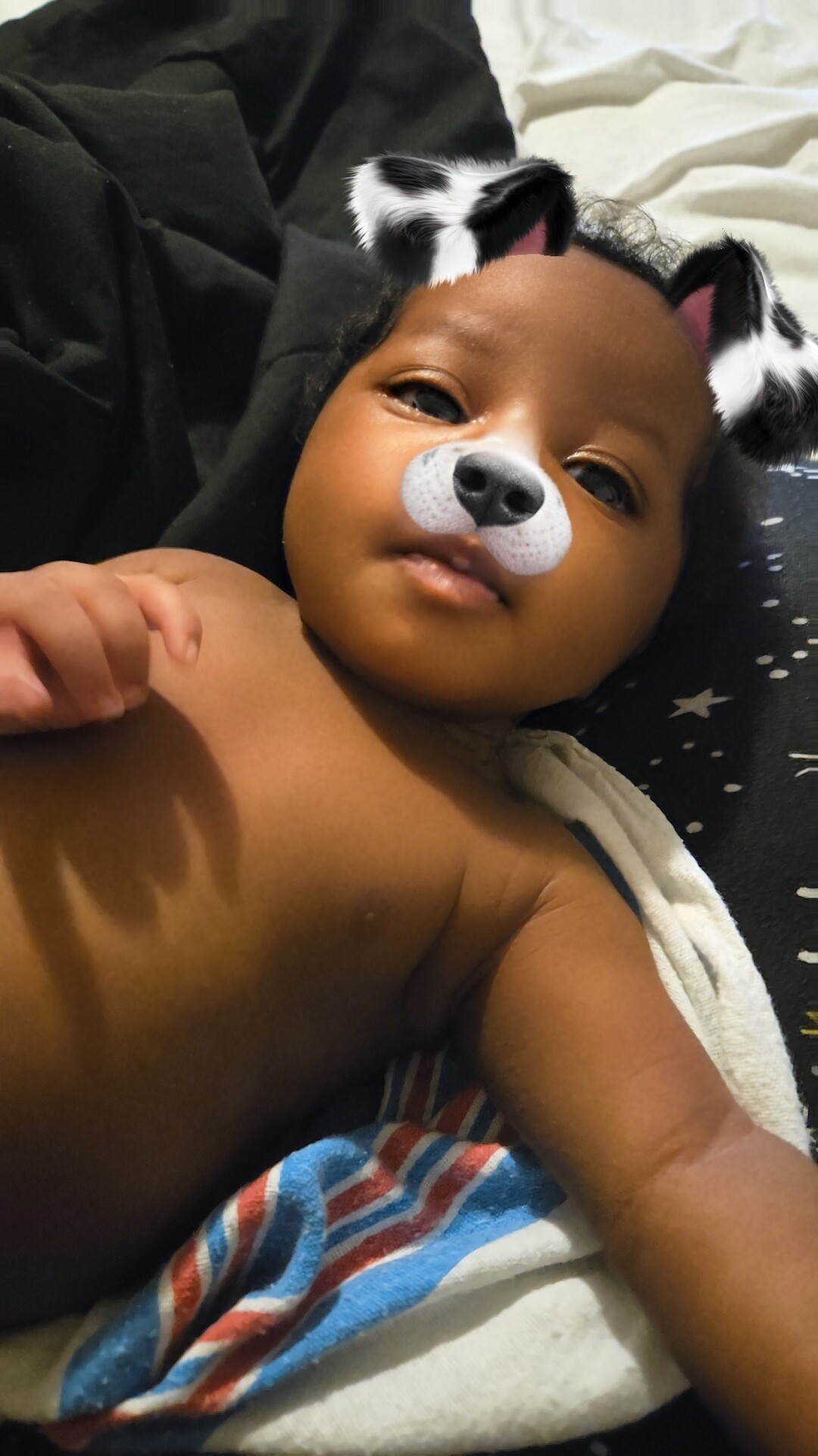 Baby lying down with a playful dog filter (black and white ears and a gray nose) over the face.
