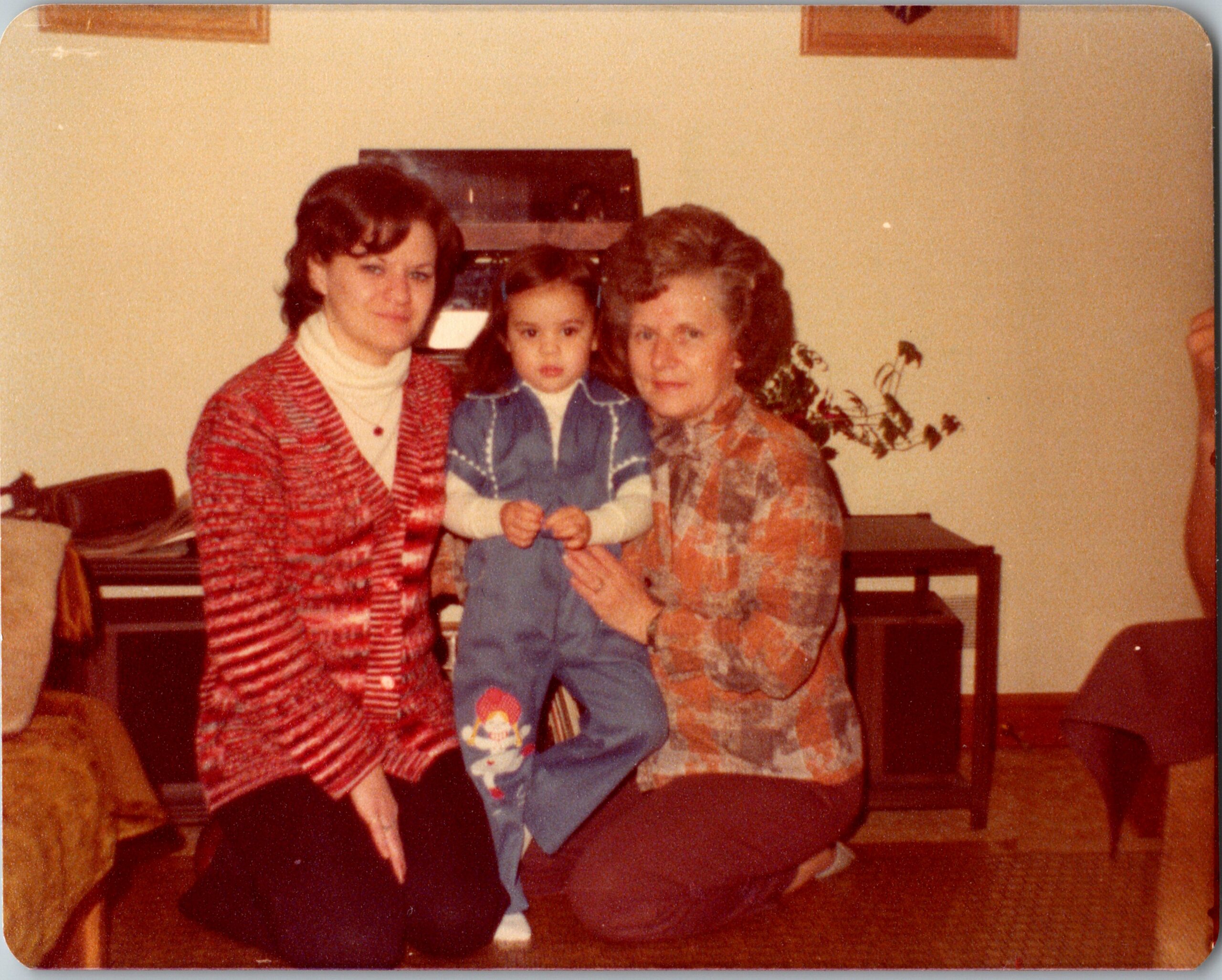 Three people posing in a living room: two women with a child in blue overalls.
