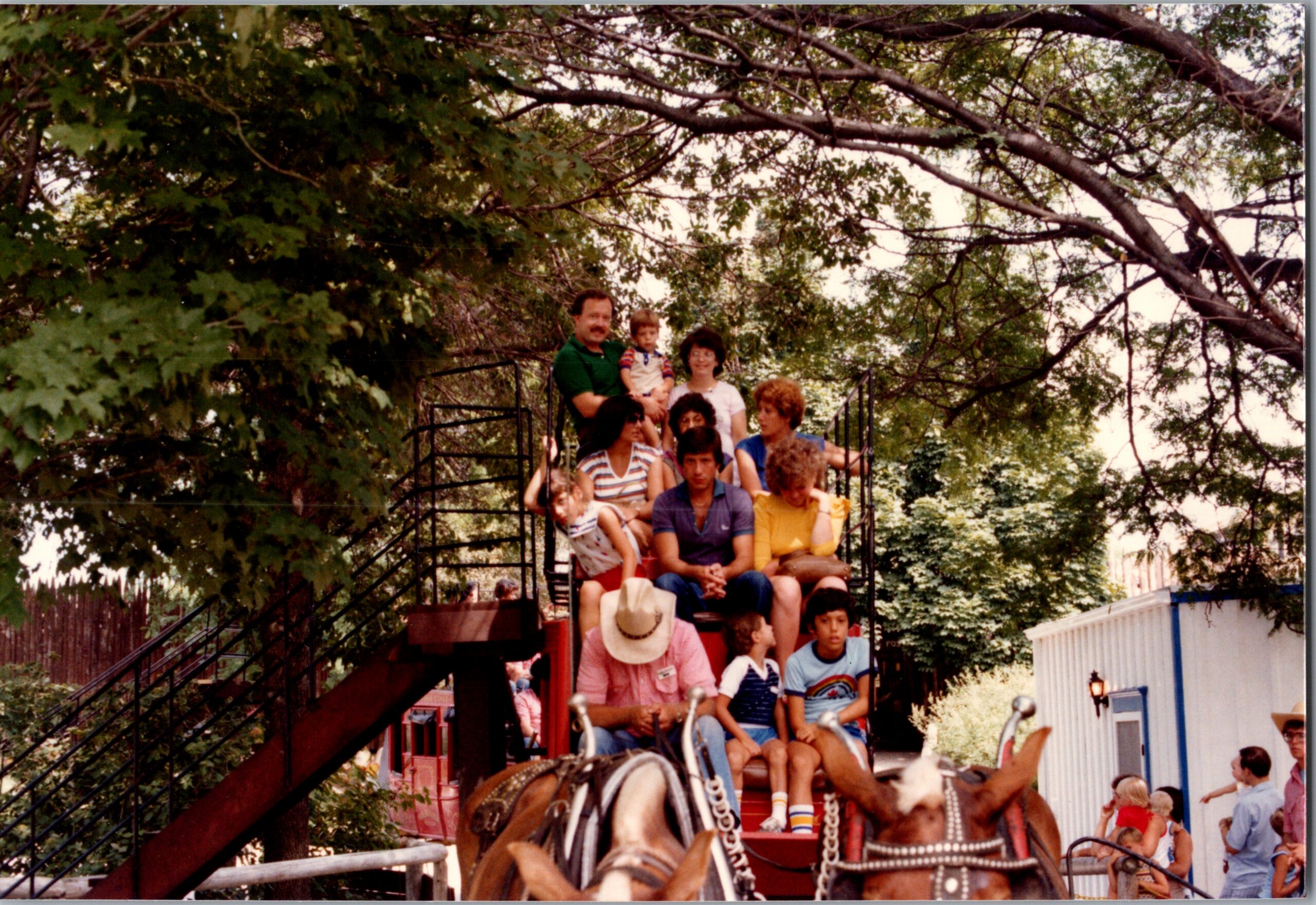 Family members ride a horse-themed amusement ride on a sunny day among green trees and a fence backdrop.