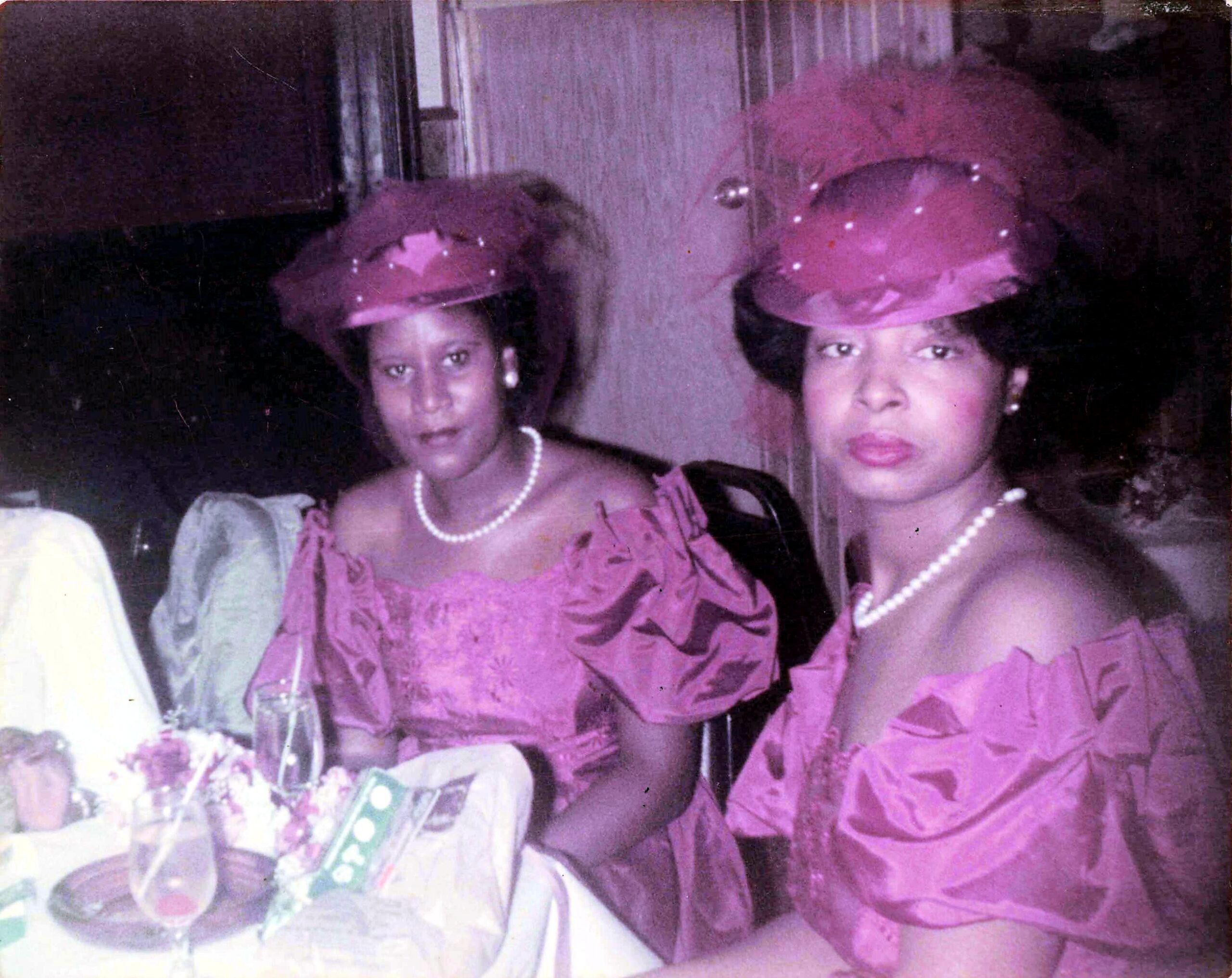 Two women in matching pink outfits and hats with pearl accessories pose together at a formal event, one wearing a pearl necklace and the other a string of pearls near a table with decorations and drinks in the foreground.