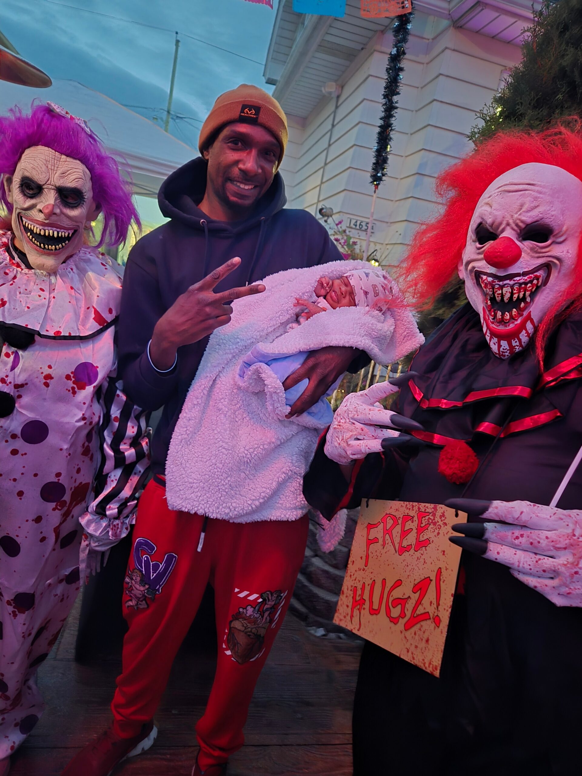 Two scary clowns stand beside a smiling man who cradles a newborn wrapped in a white blanket outside a house; a 'FREE HUGS!' sign is held nearby.