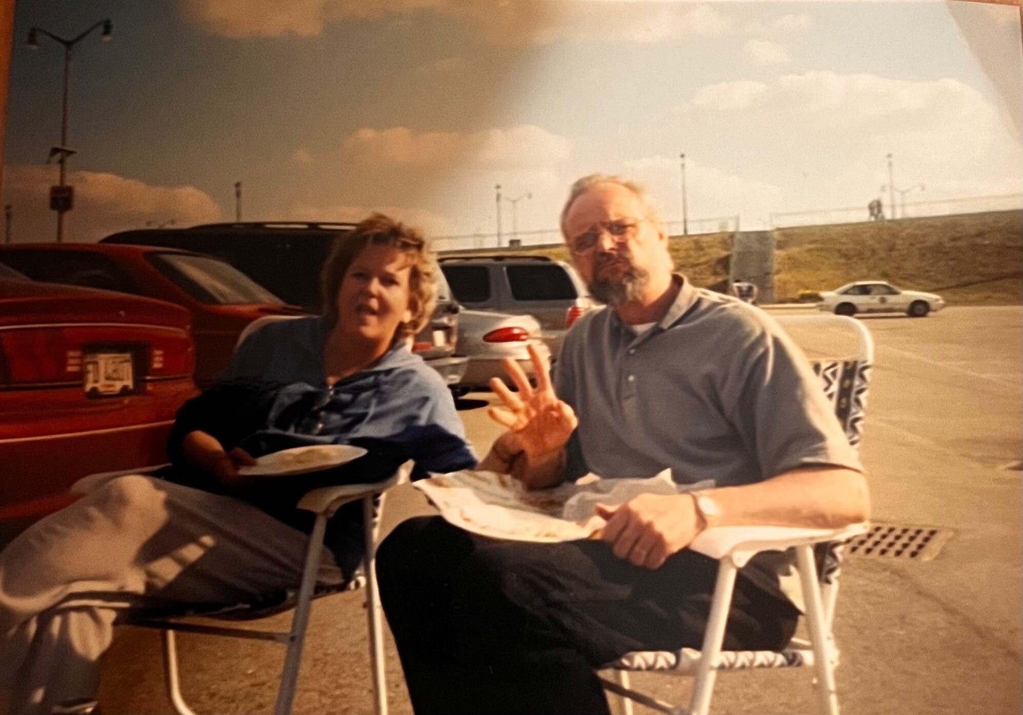 Two adults sit in folding chairs at a table in a parking lot, eating takeout as cars sit in the background.