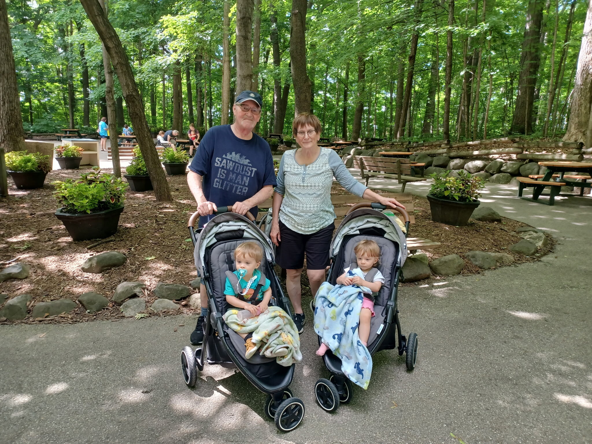Older couple poses with two double baby strollers in a wooded park; children wrapped in blankets in front.