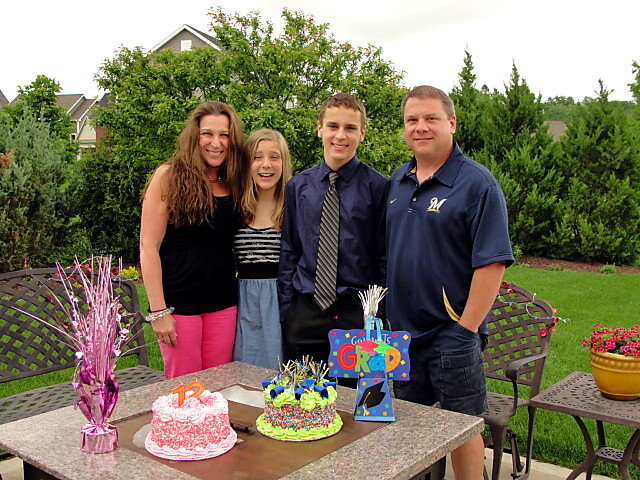 Family of four smiling behind a table with two decorated graduation cakes in a backyard celebration.