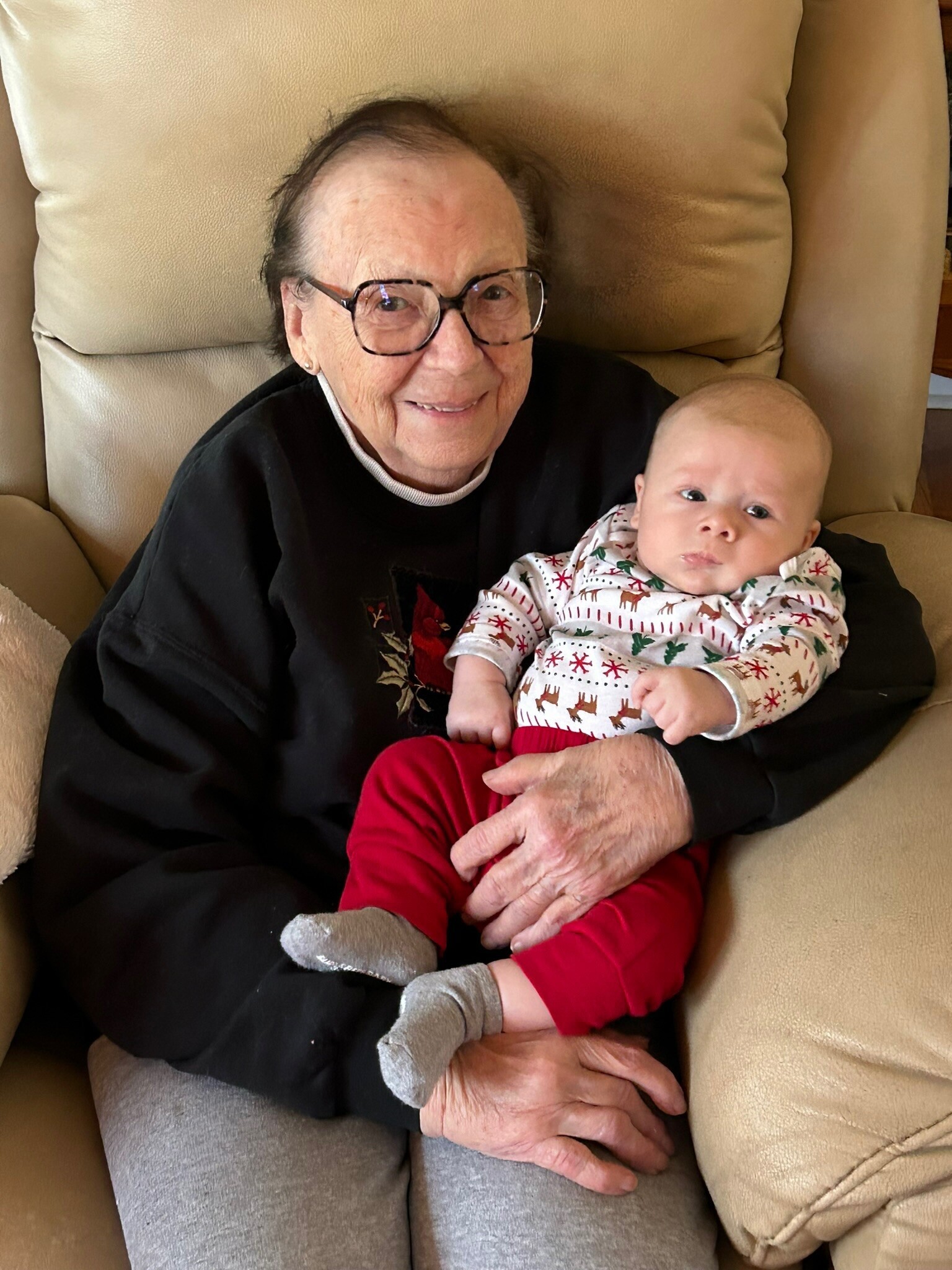 Older woman with glasses smiling while sitting on a beige couch and holding a baby.