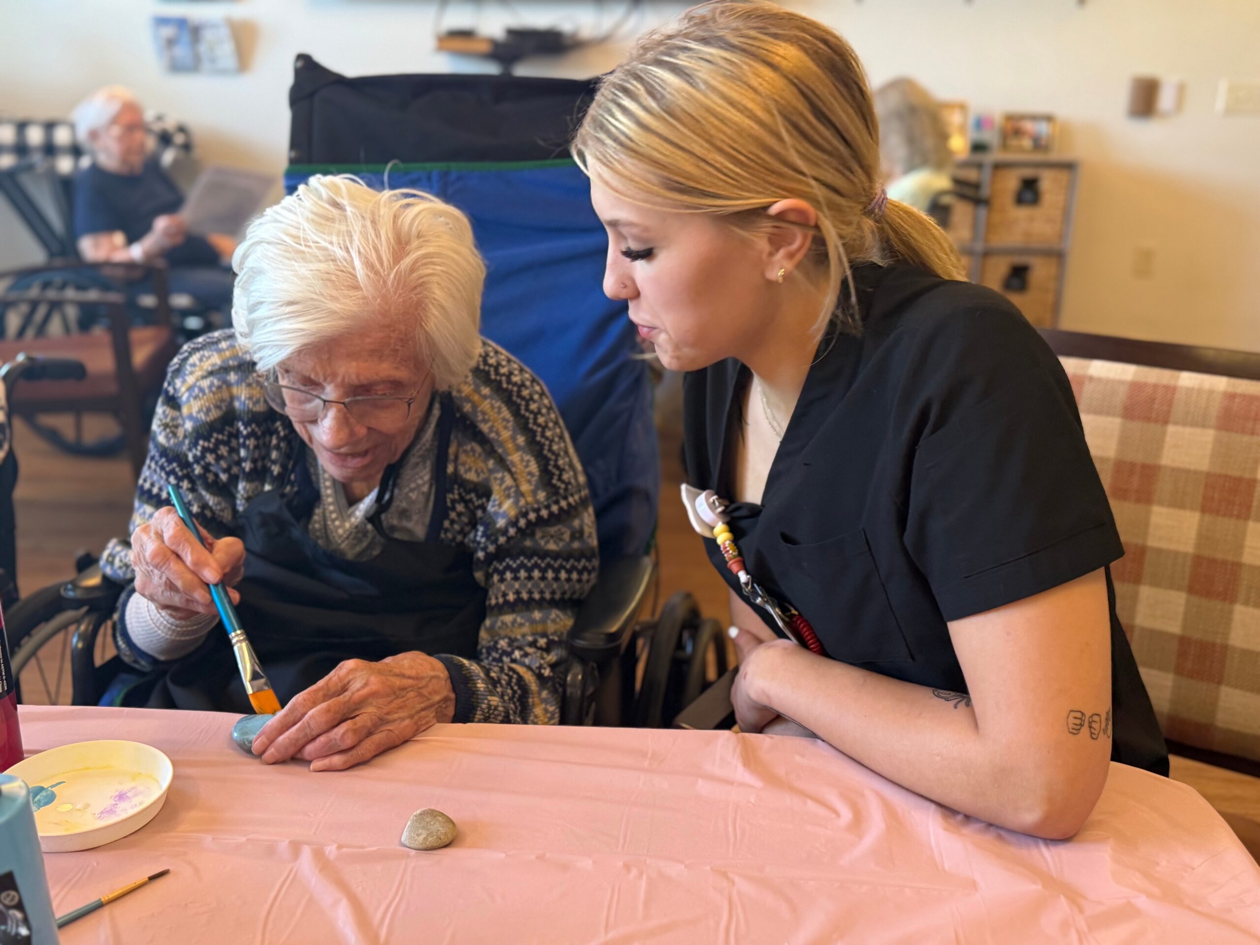 Elderly woman in a wheelchair painting at a table with a caregiver leaning in to help, pastel pink tablecloth and art supplies visible.