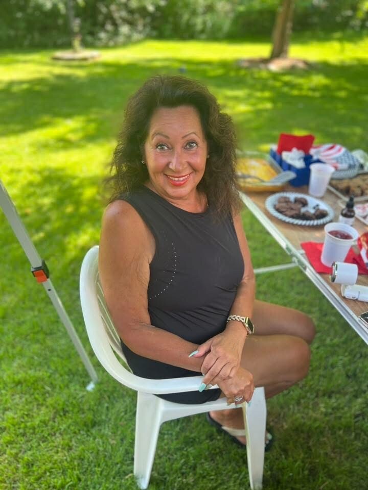 Smiling woman in a black sleeveless dress sits on a white plastic chair at a sunny outdoor picnic with a table of food beside her
