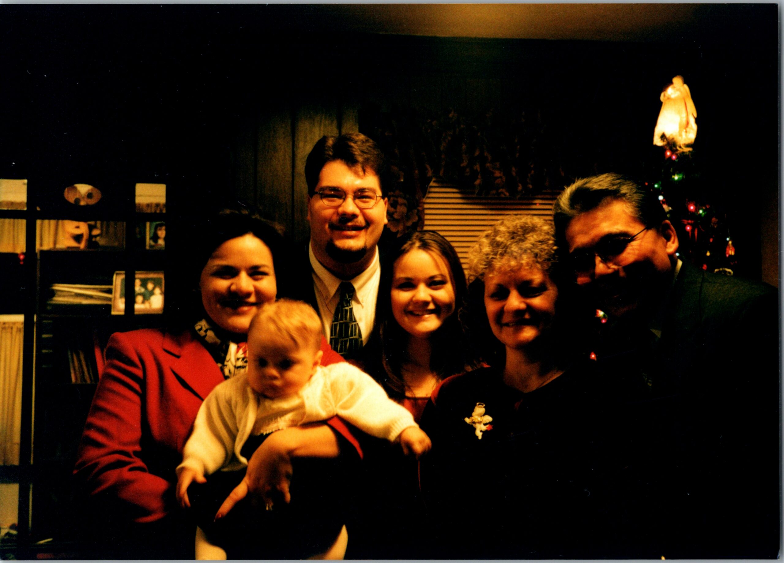 A group of smiling adults posing indoors with a baby, Christmas tree visible in the background.