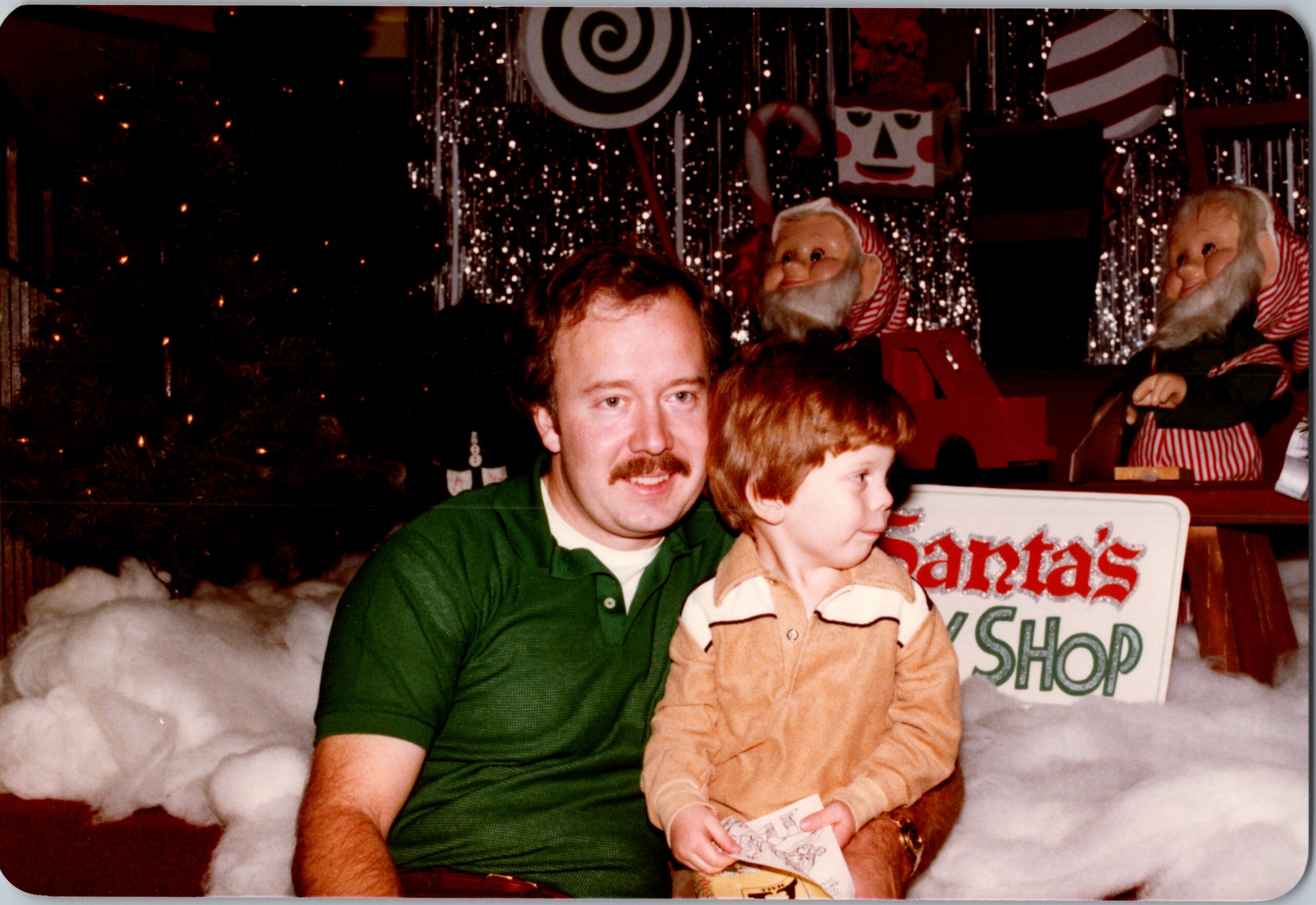 Man with a mustache in a green polo sits with a young boy on a snowy, festive backdrop for Christmas photos.