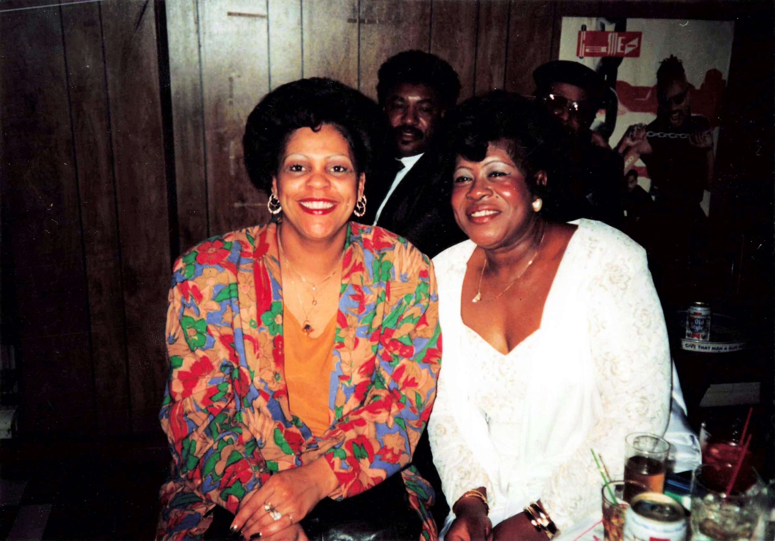 Two women smiling at a party, one in a bright floral blazer and the other in a white lace dress, posed at the camera.