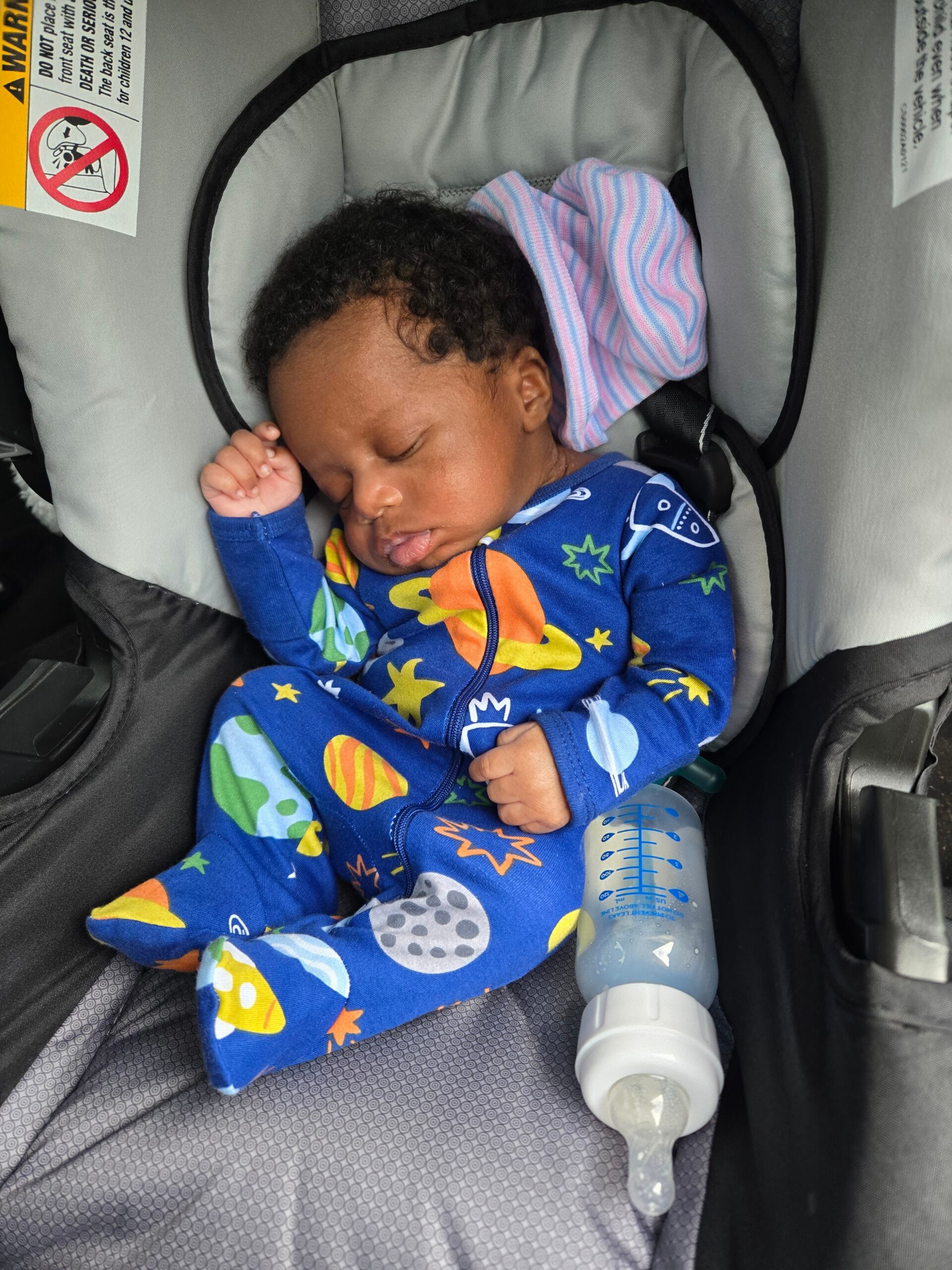 Infant sleeping in a car seat wearing a blue space-themed onesie, with a pink-and-blue striped blanket nearby and a bottle resting beside.