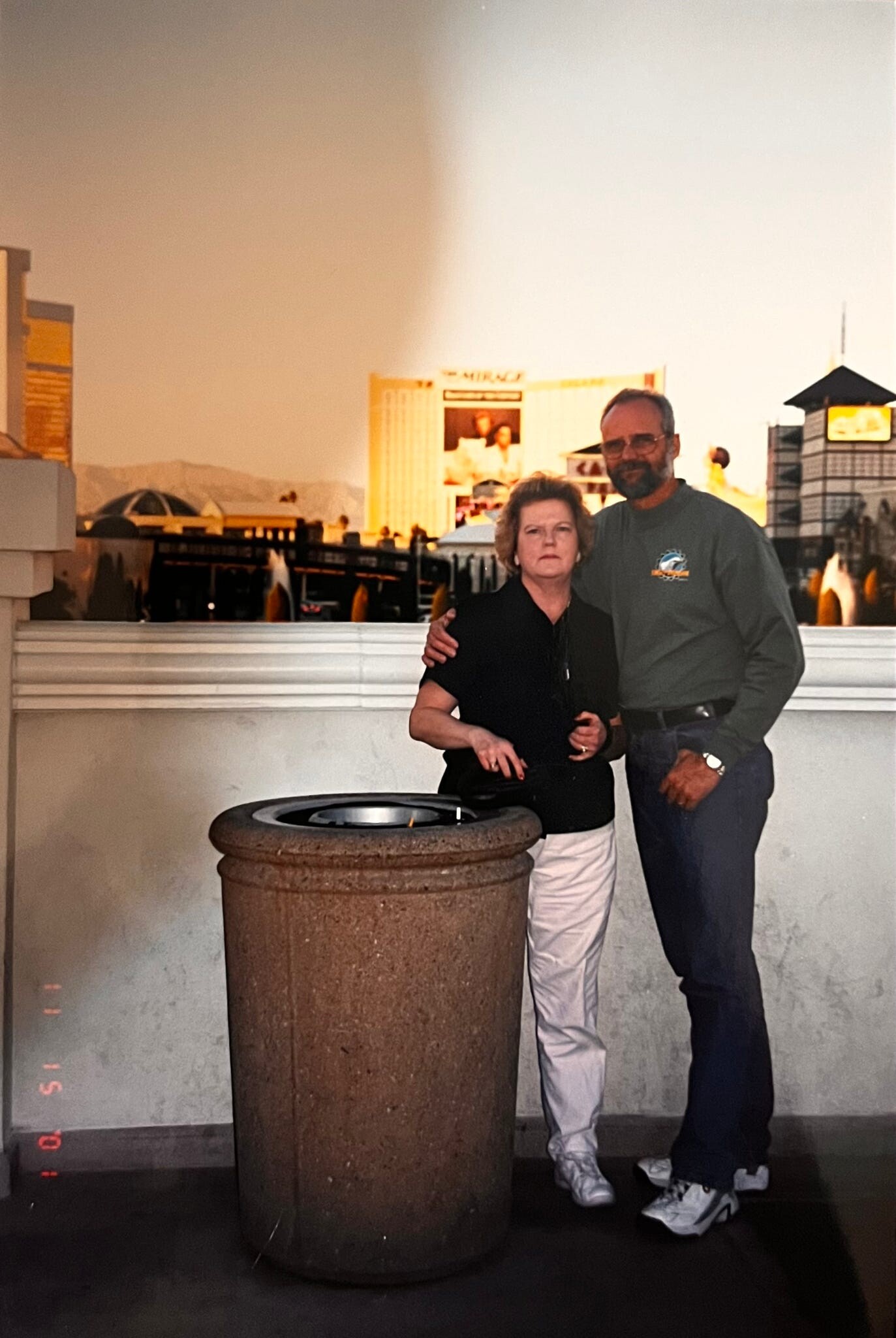 Two adults posing together beside a large brown trash can in an indoor shopping area, looking at the camera.