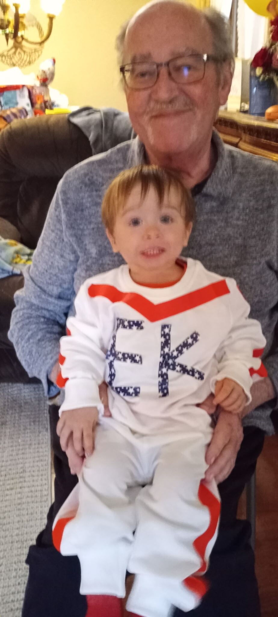 Grandfather with glasses sitting on a couch, holding a smiling toddler in a white outfit with red stripes.