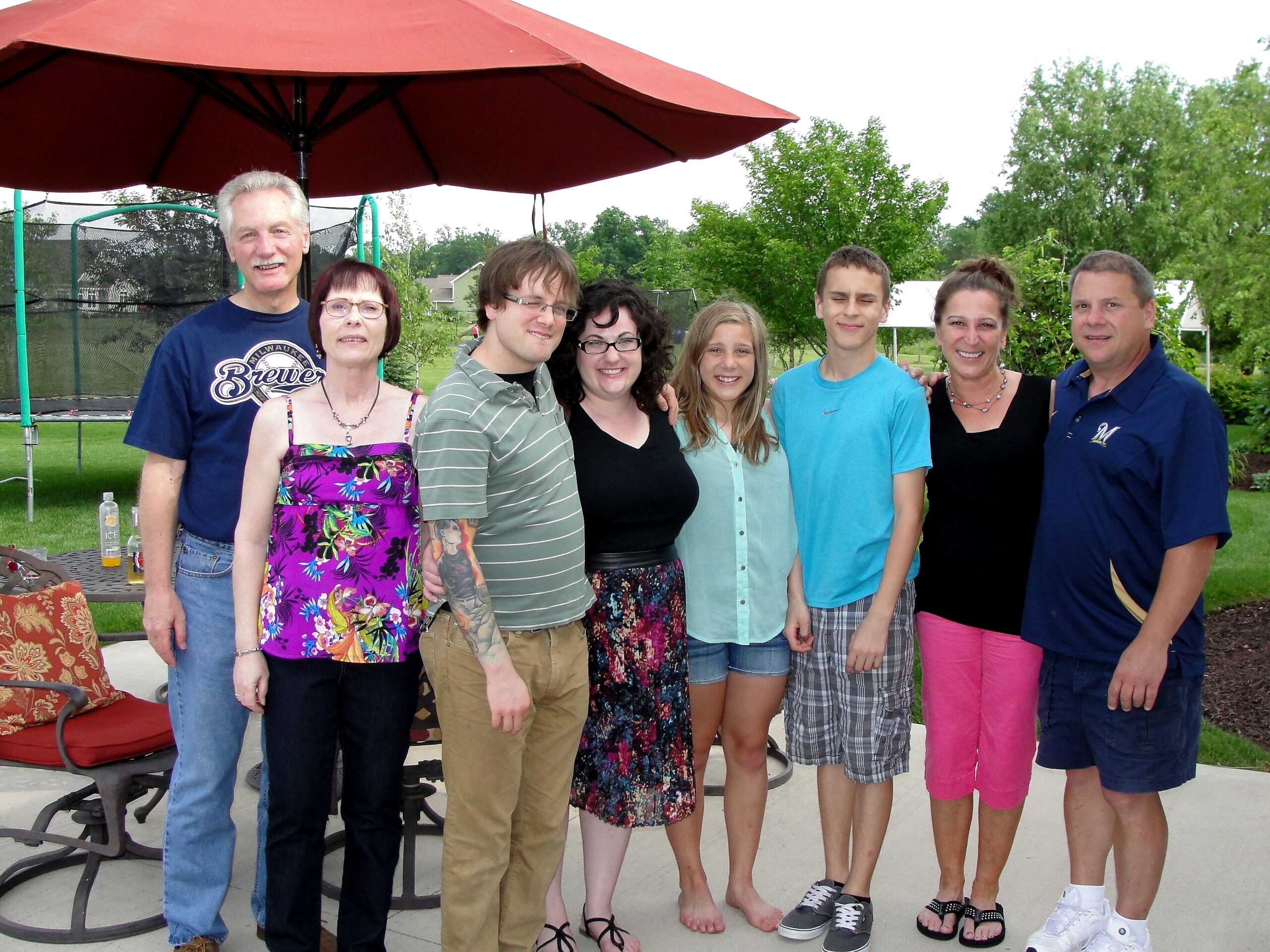Group of nine people posing together outdoors under a large red umbrella, smiling at the camera in a yard with trees in the background.