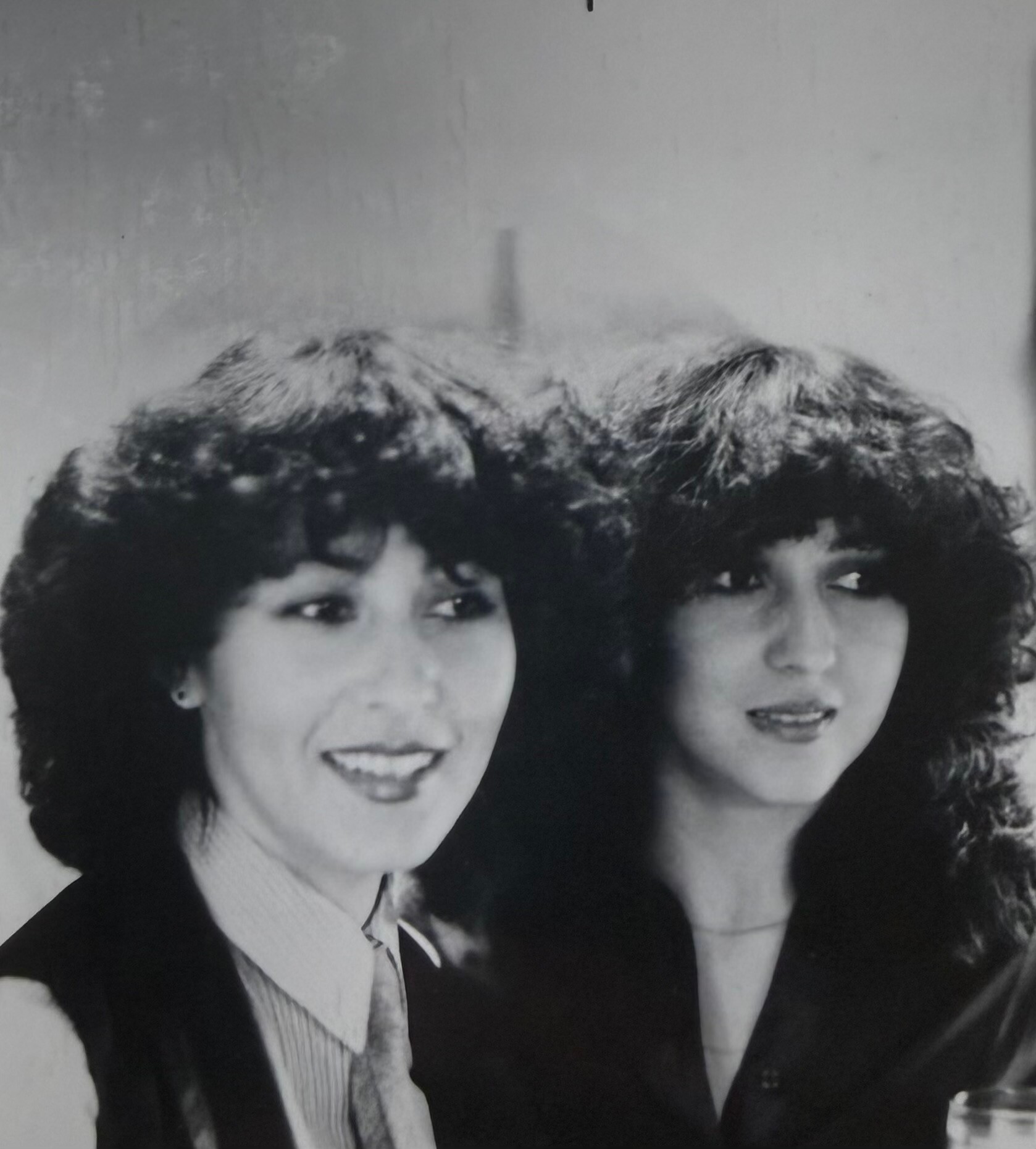 Two women pose closely for a black-and-white portrait, smiling at the camera, wearing blouses and dark vests/jackets.