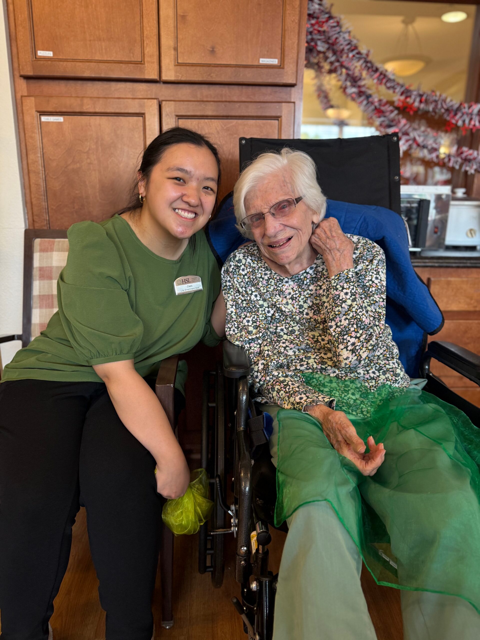 Young caregiver and elderly woman sit together, smiling in a wheelchair beside wood cabinets and festive garlands.