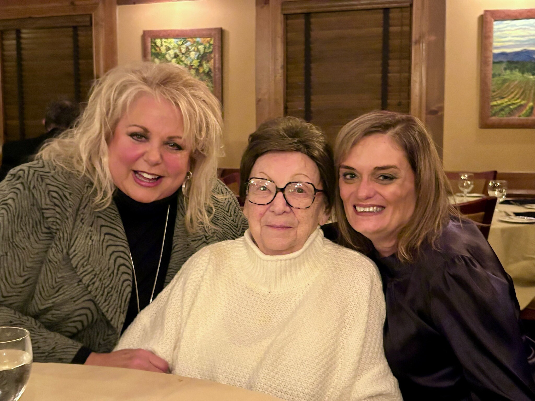 Three women smiling at a restaurant table, taking a selfie together in warm lighting.