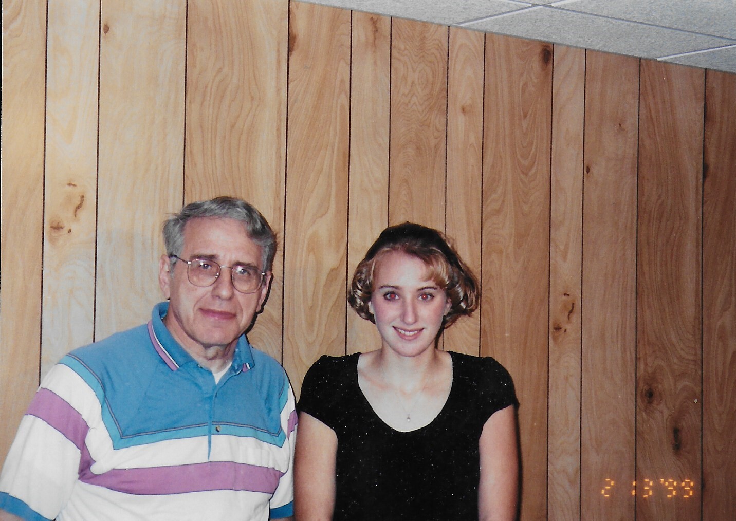 Older man with gray hair and glasses standing beside a young woman in a sparkly black top, against a vertical wood-panel wall.