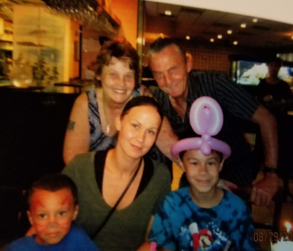 Family group posing for a photo at a party: a woman seated with two kids in front, one wearing a pink balloon hat and the other with face paint, with two adults behind them smiling.
