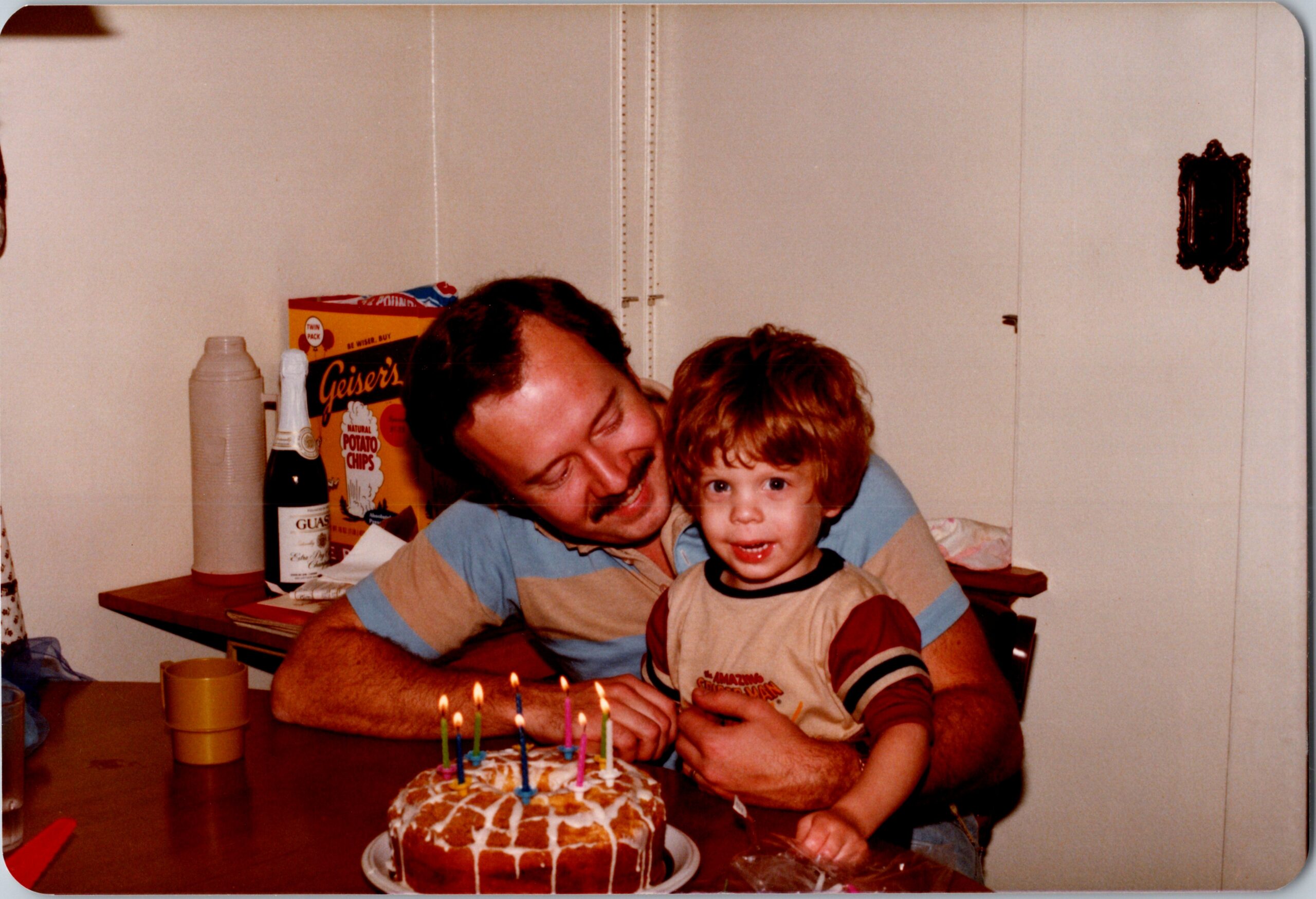 Man cuddles a child at a birthday cake with lit candles on a kitchen table.
