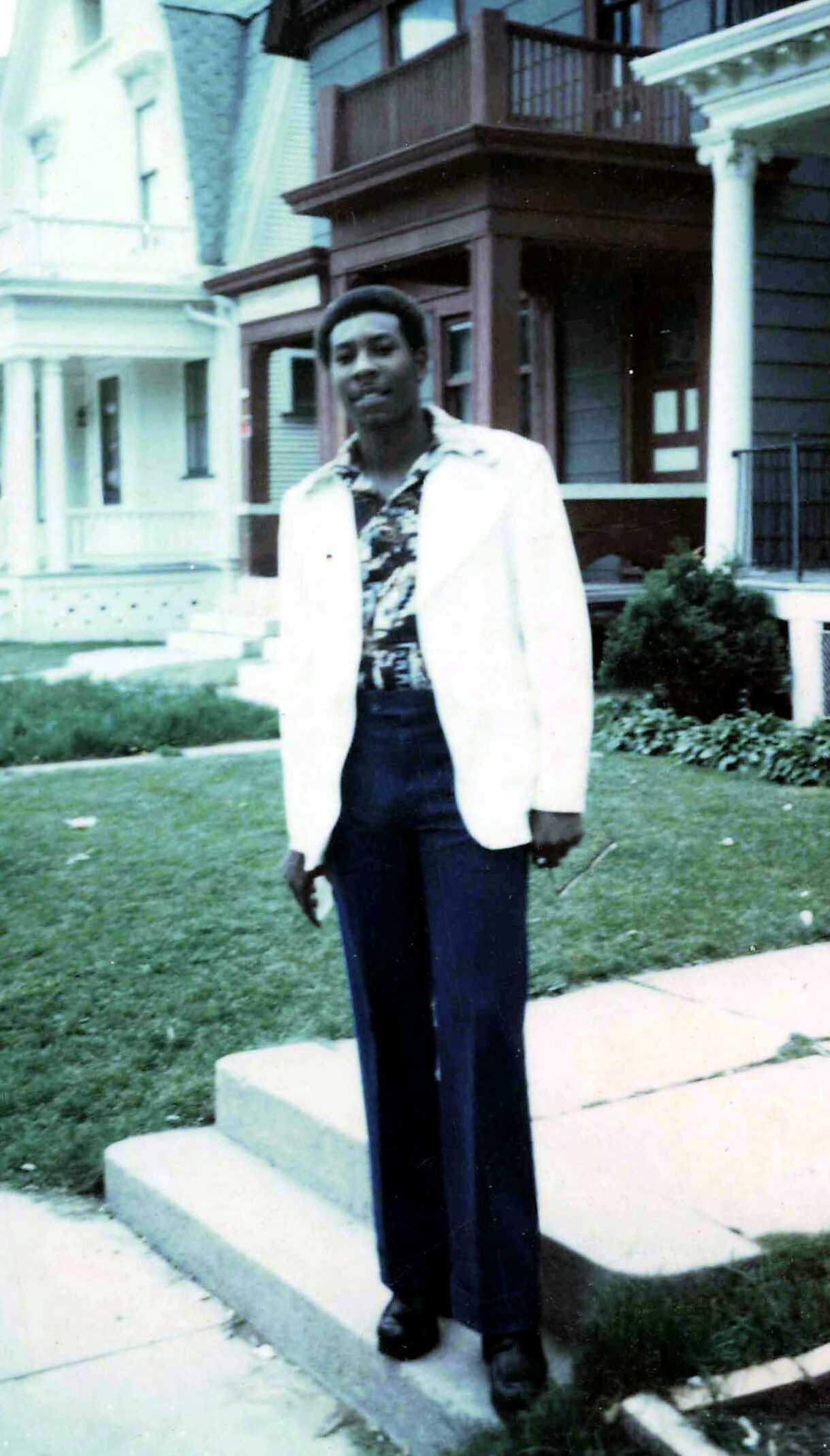 Young man in a white blazer and patterned shirt stands on stone steps in a residential street.