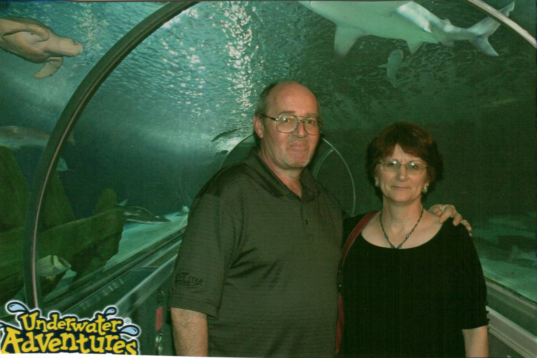 Couple posing in an underwater tunnel with sharks swimming overhead, logo 'Underwater Adventures' visible at corner