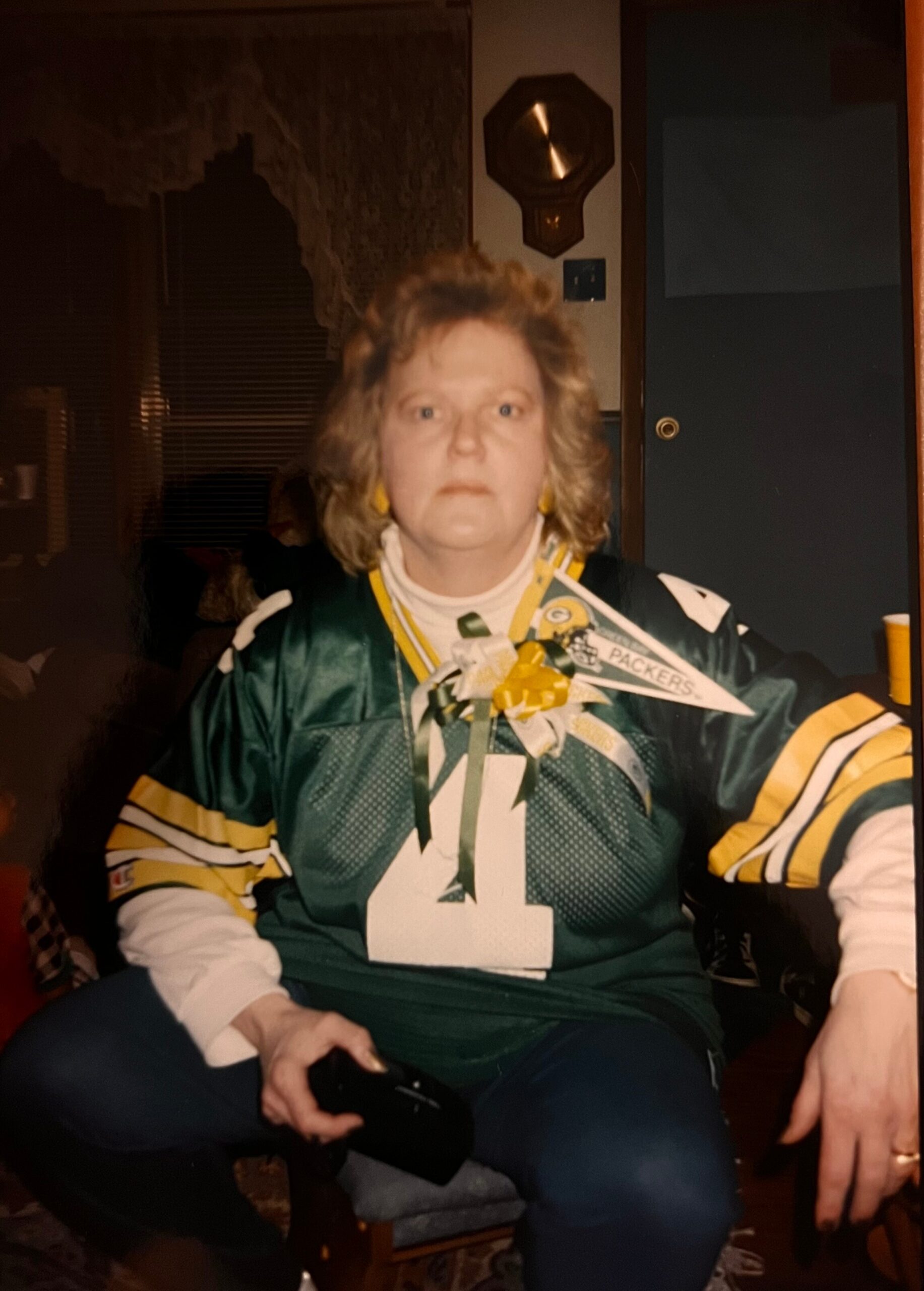 Older woman with short blonde hair wearing a Green Bay Packers jersey, seated indoors, holding a black object in her right hand.