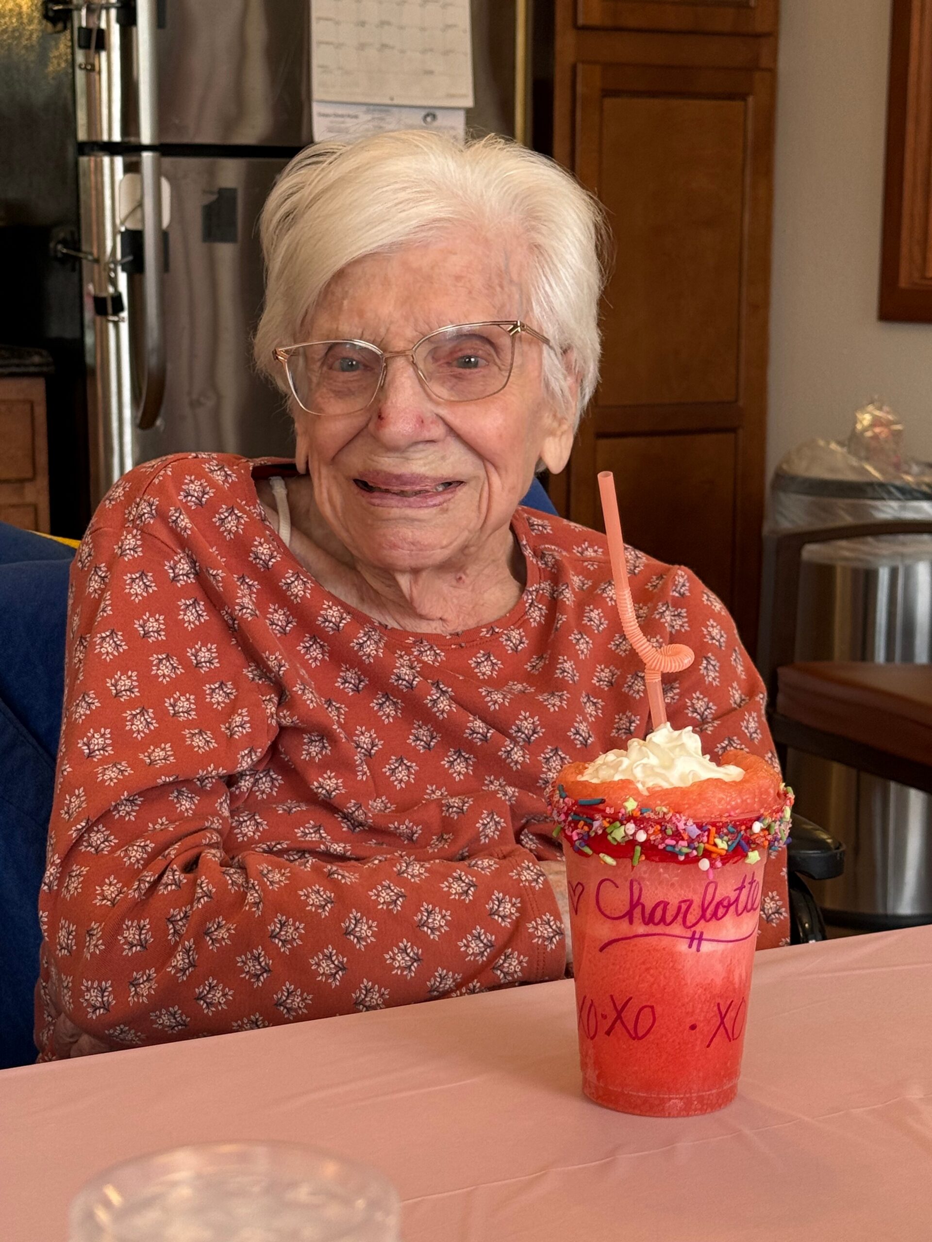 Smiling elderly woman with white hair and glasses sits at a pink table with a tall pink celebratory milkshake topped with whipped cream and colorful sprinkles in front of her in a kitchen setting.