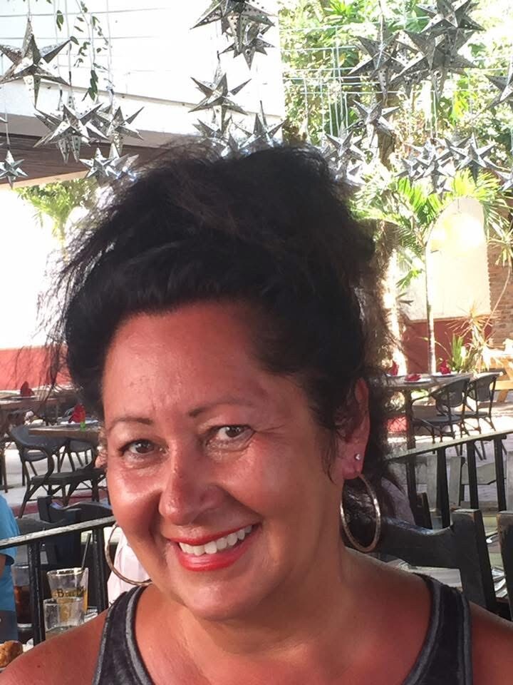 Smiling woman with dark hair in a ponytail wearing hoop earrings at an outdoor cafe.