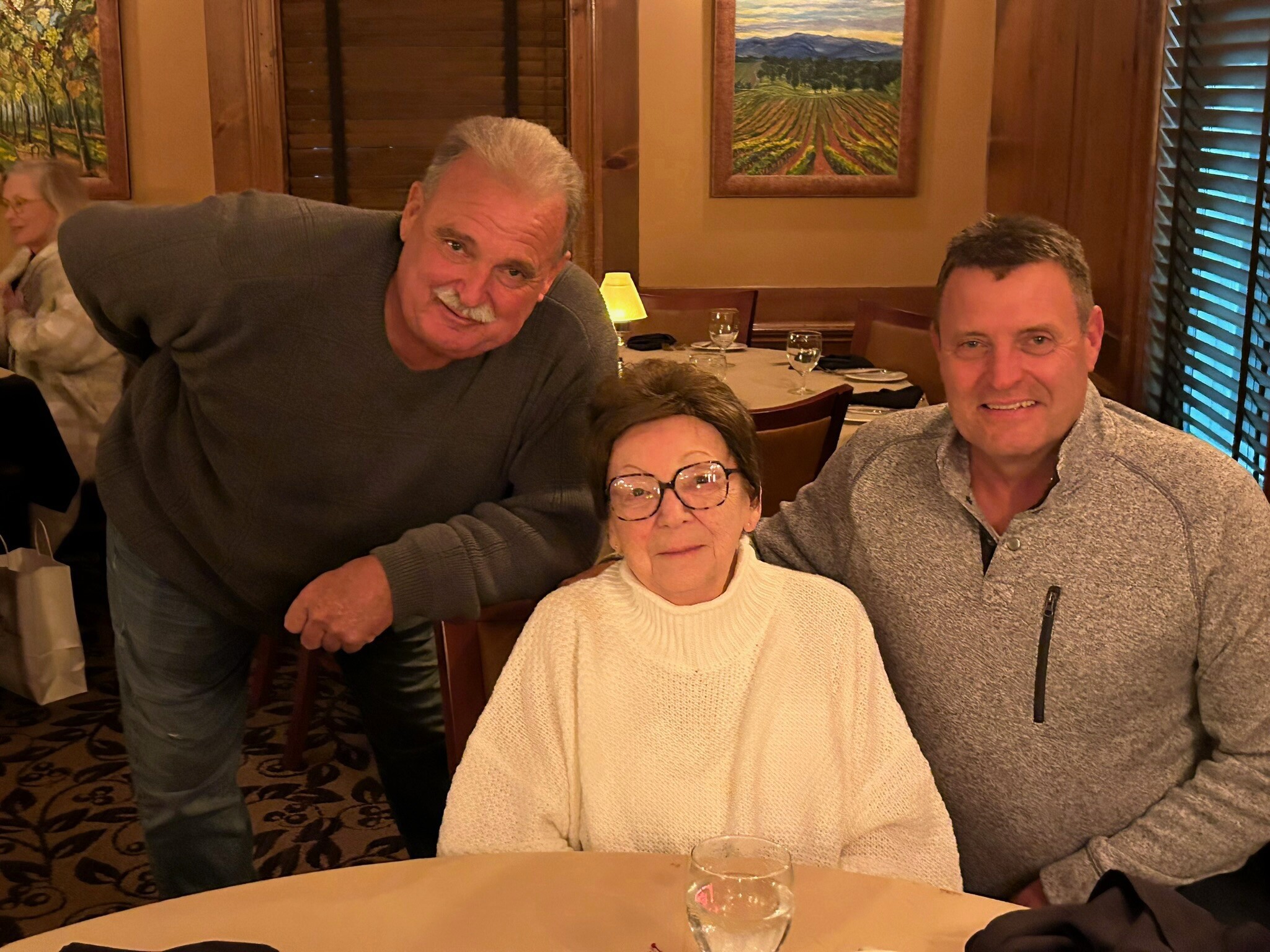 Three adults dining together at a restaurant table: a woman in a white sweater center, a man leaning from the left, and a man in a gray knit on the right, with glasses and dinnerware on the table behind them.