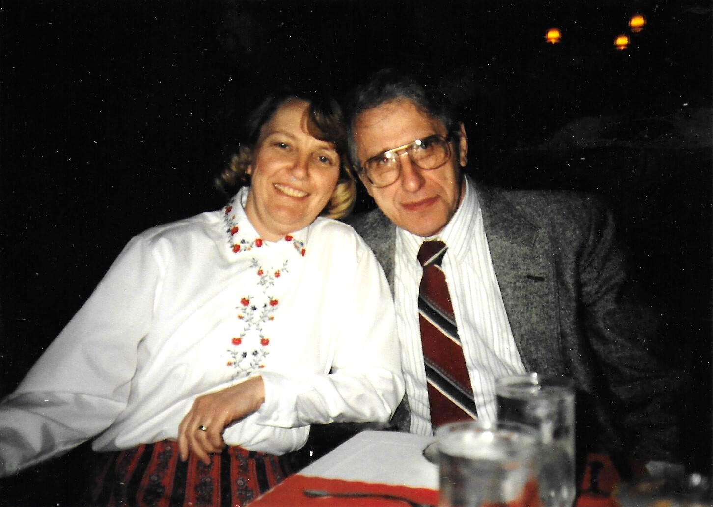 Smiling couple seated at a dinner table in a dim setting; woman wears a white embroidered blouse and the man wears a gray jacket and striped shirt, leaning together.