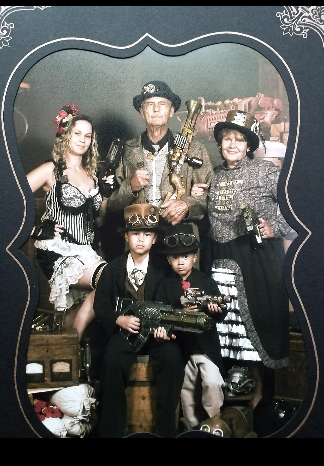 Family in steampunk costumes posing for a portrait, with goggles and brass gear, holding prop weapons while a backdrop of gears looms behind them.