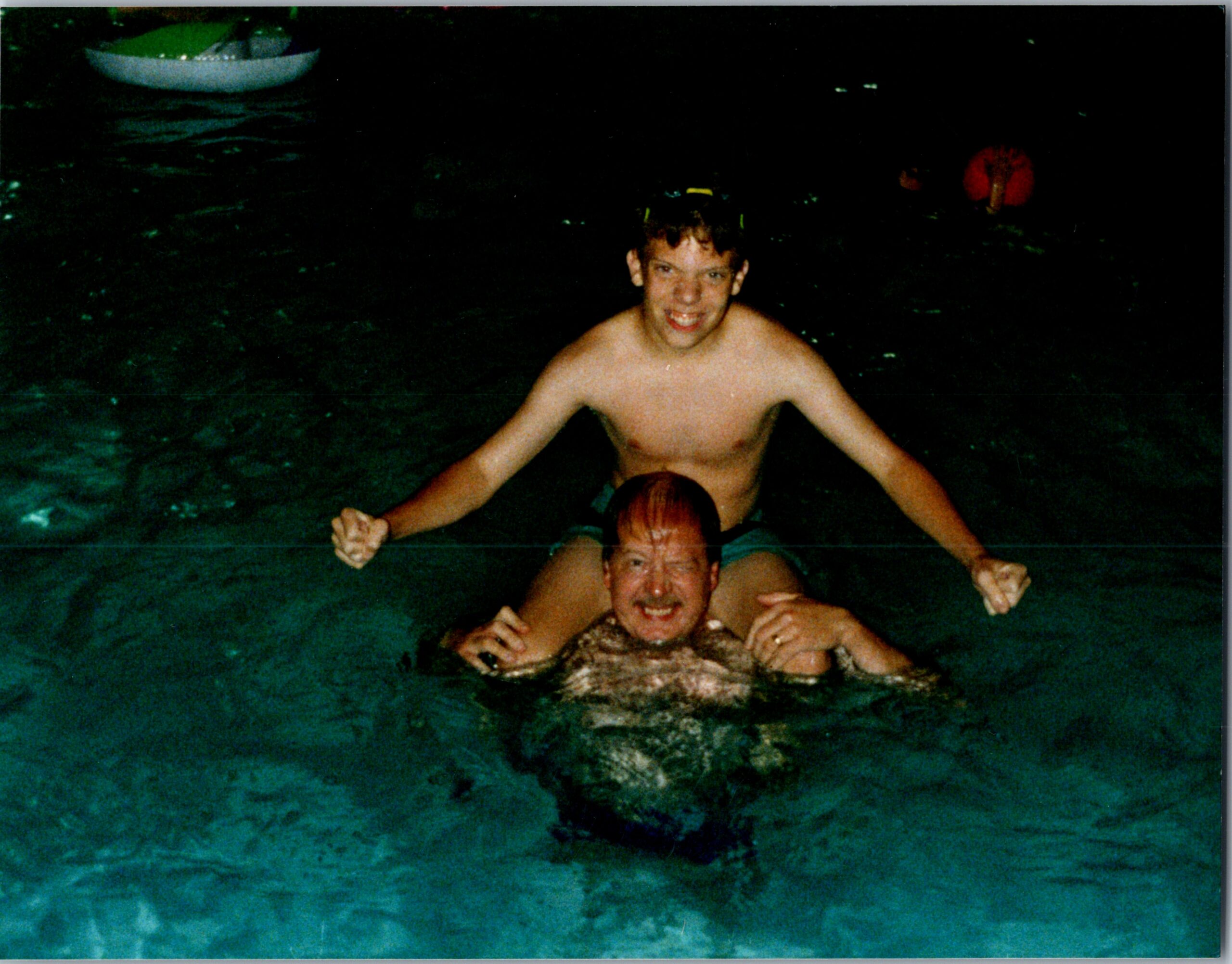Two people swimming in dark water at night, one person sitting on top of the other and smiling, with a small boat in the background left.