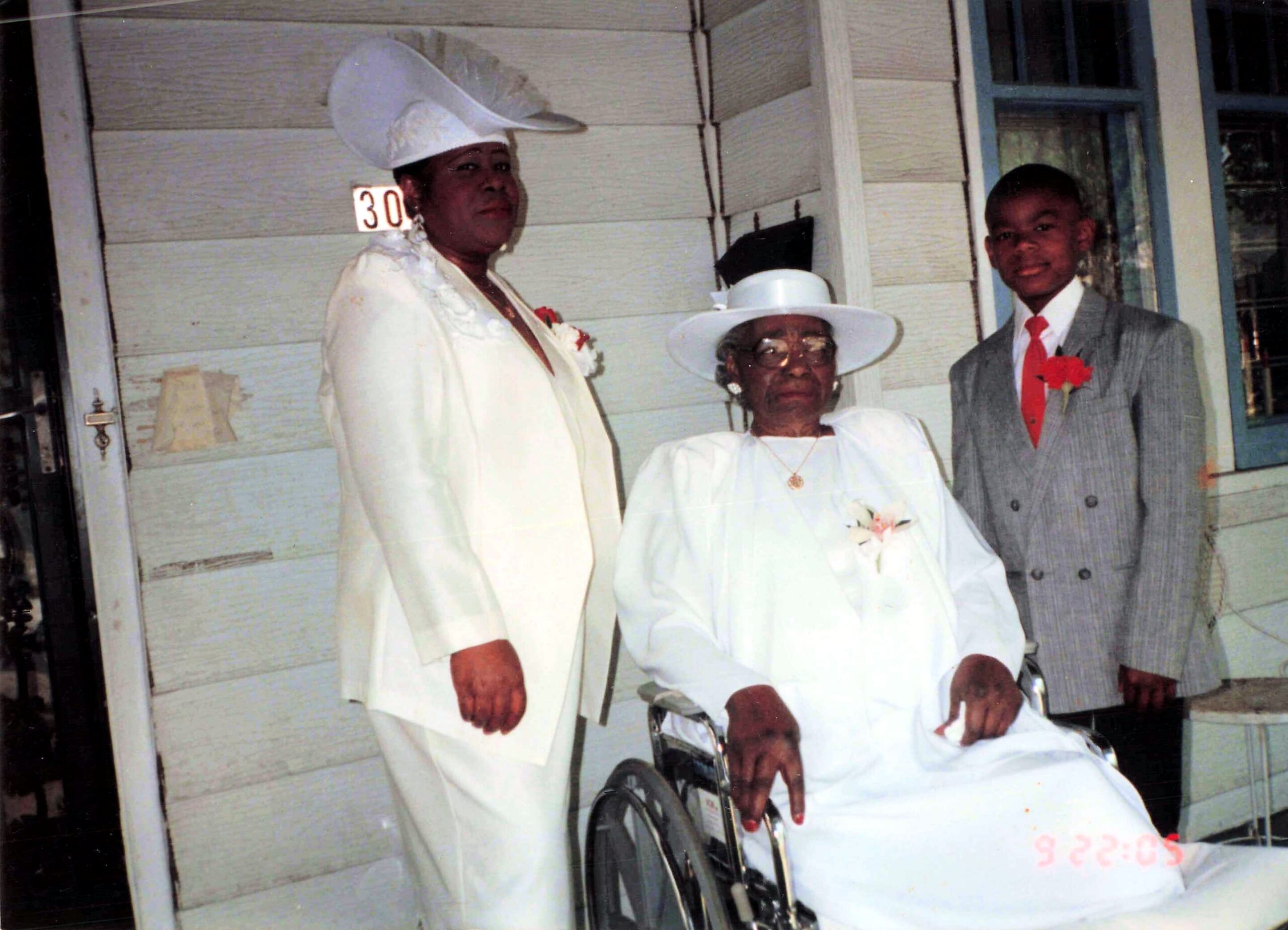 Three people in formal dress pose outside a house; an elderly woman in a wheelchair flanked by two others wearing hats and corsages, house number 30 visible.