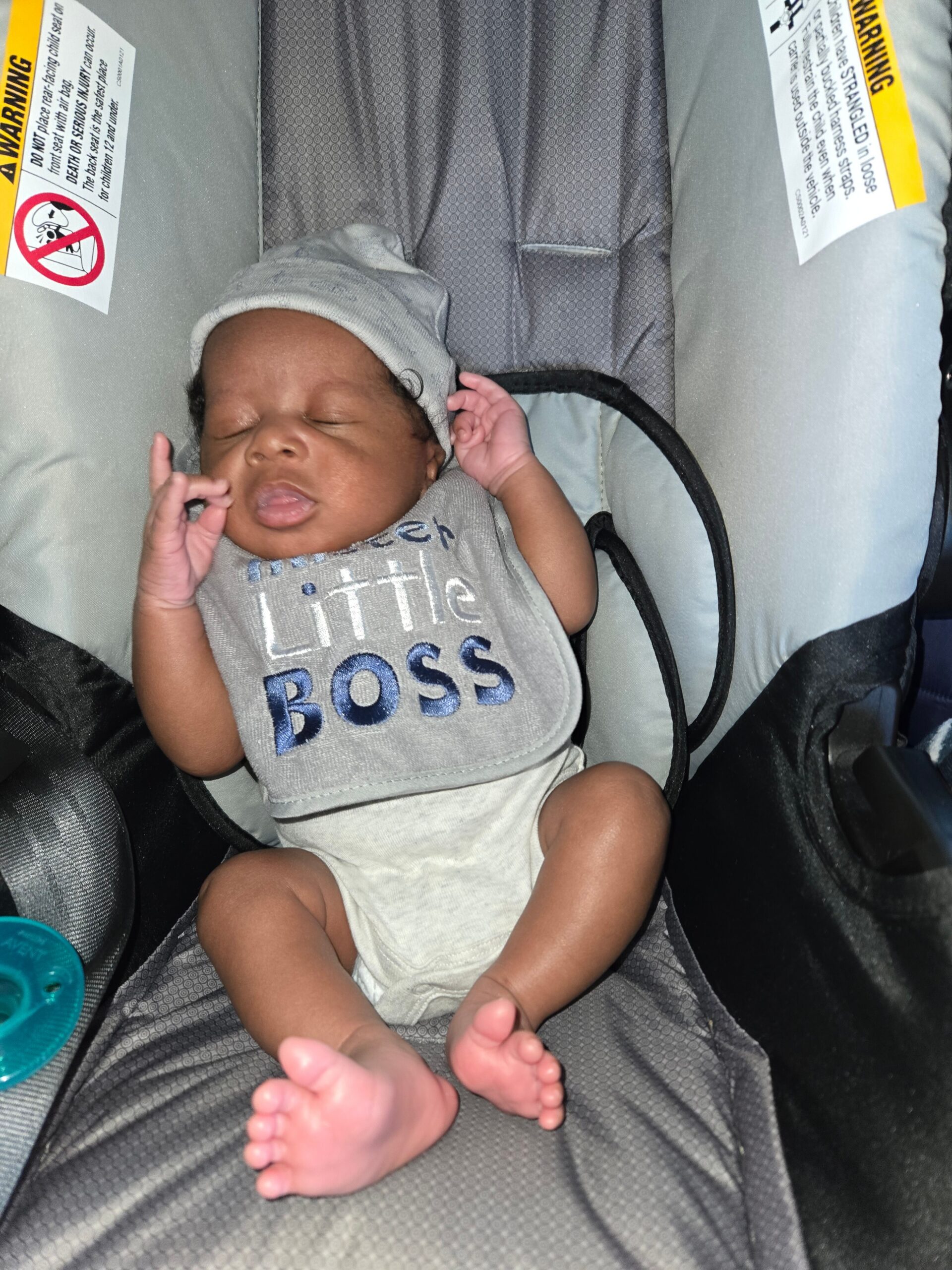 Sleeping baby in a gray car seat, wearing a light gray hat and a onesie that reads 'little boss'.