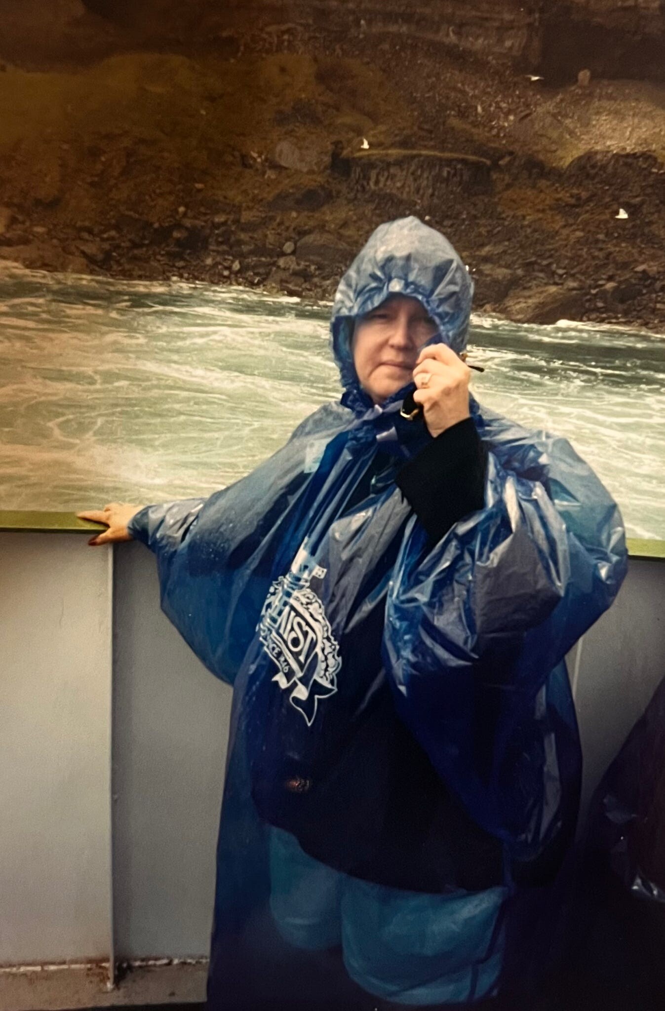 Person in a blue rain poncho with hood, standing at a railing by rough ocean water.
