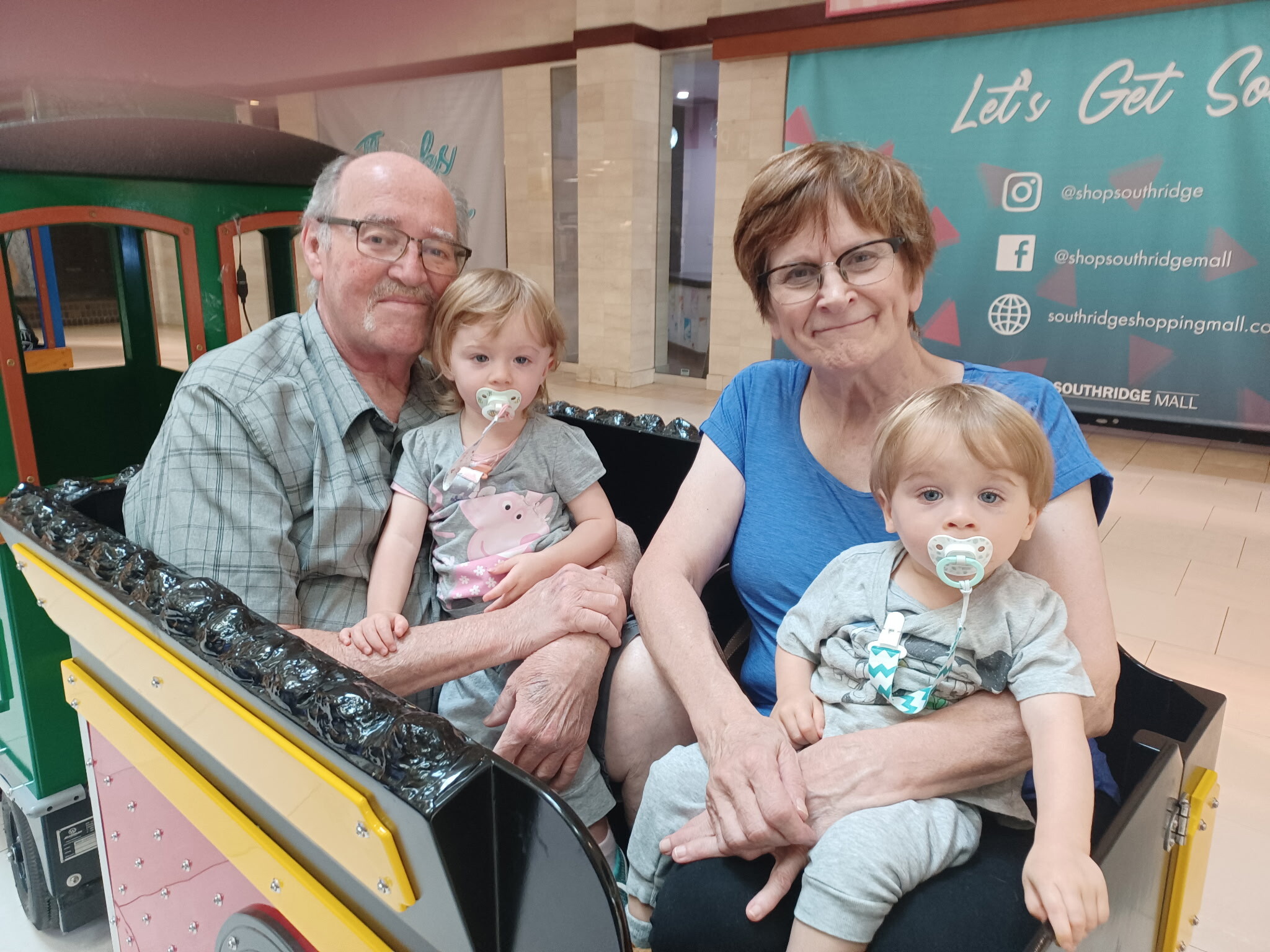 Grandfather and grandmother with two young children posing on a colorful kiddie ride inside a shopping mall.
