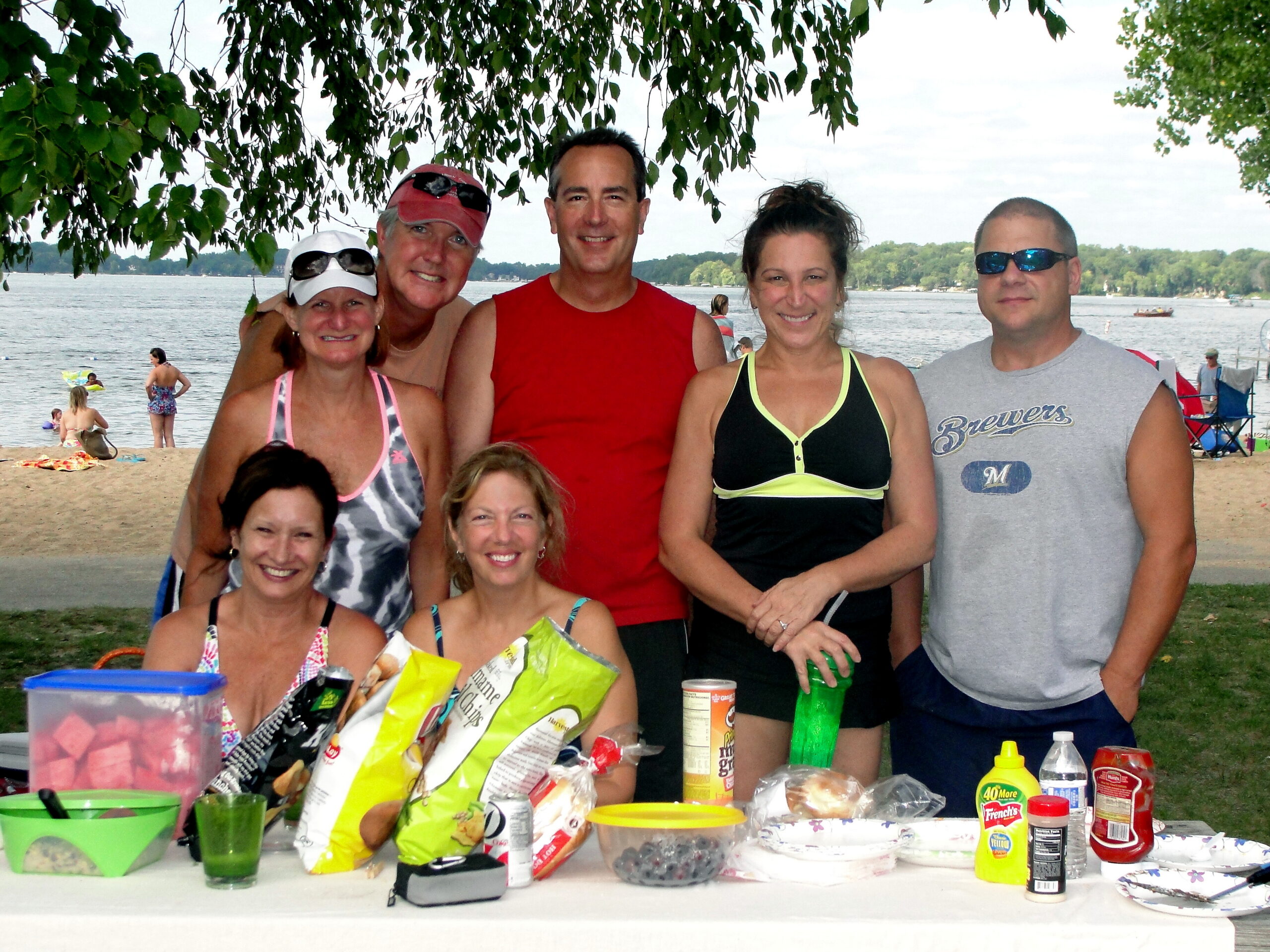 Group of adults at a lakeside picnic posing for a photo, with snacks and drinks on a table in front. Under a tree, a beach and water in the background.