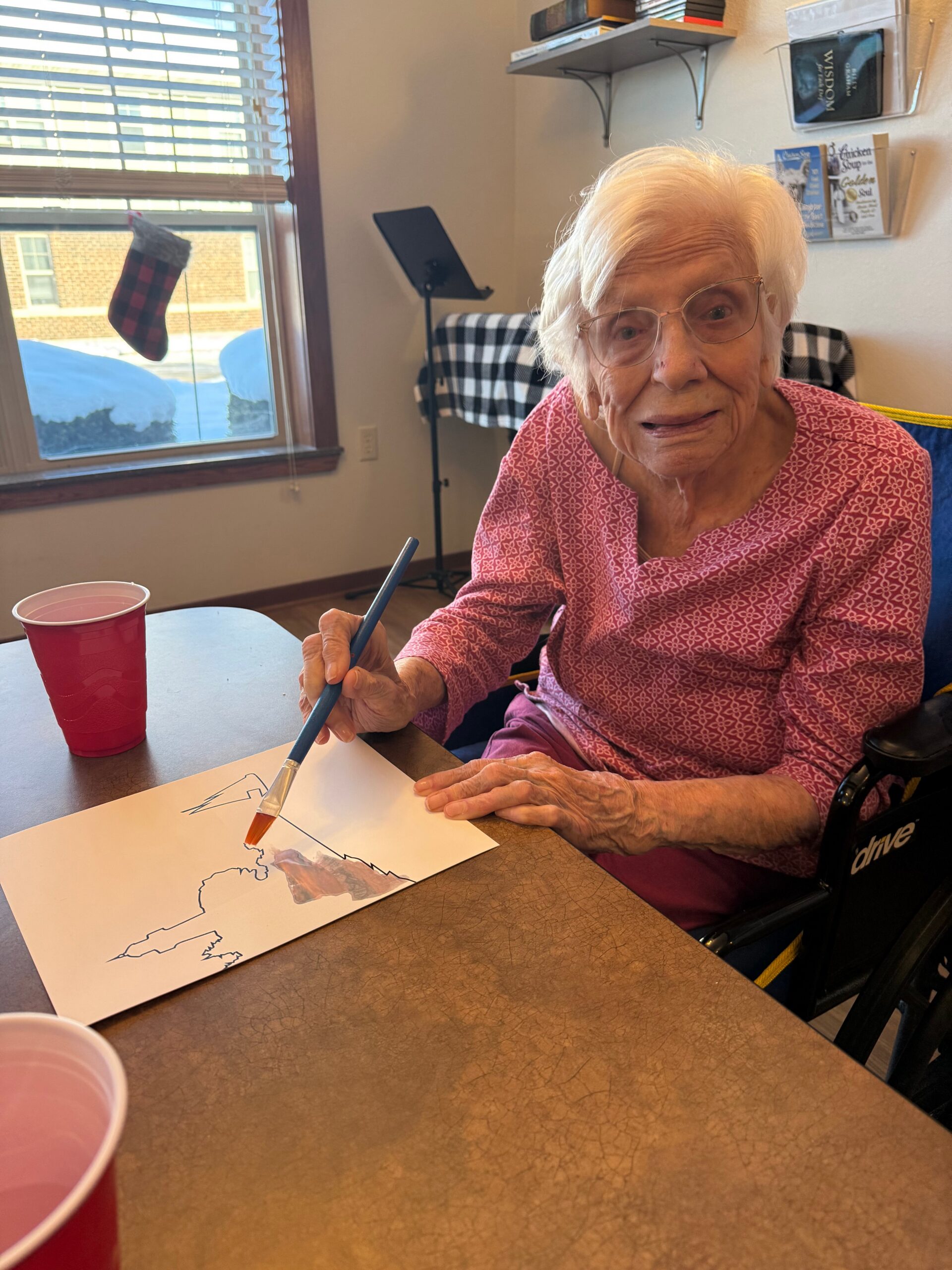 An elderly woman in a pink patterned blouse sits at a table, drawing with a blue pencil, a red cup nearby.