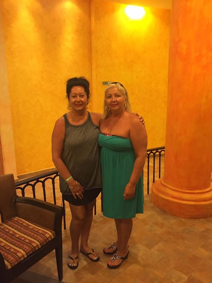 Two women posing together indoors in a warm yellow lobby, one in a gray sleeveless top and black shorts, the other in a turquoise dress with sandals.