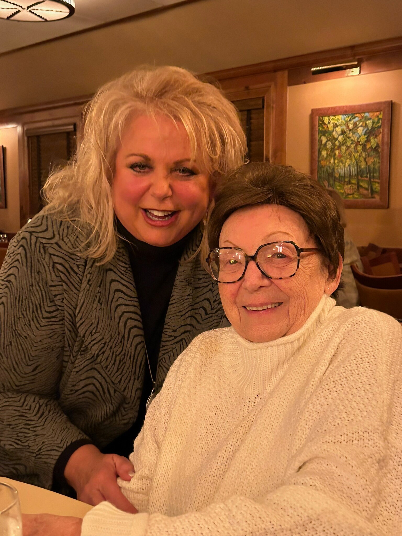 Two women smiling for a selfie at a restaurant table, one in a white knit sweater and the other in a patterned jacket with glasses in the foreground.