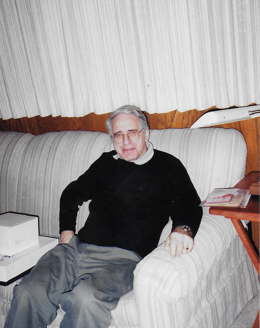 Older man with gray hair and glasses sits on a light-striped sofa, wearing a black sweater and gray pants.